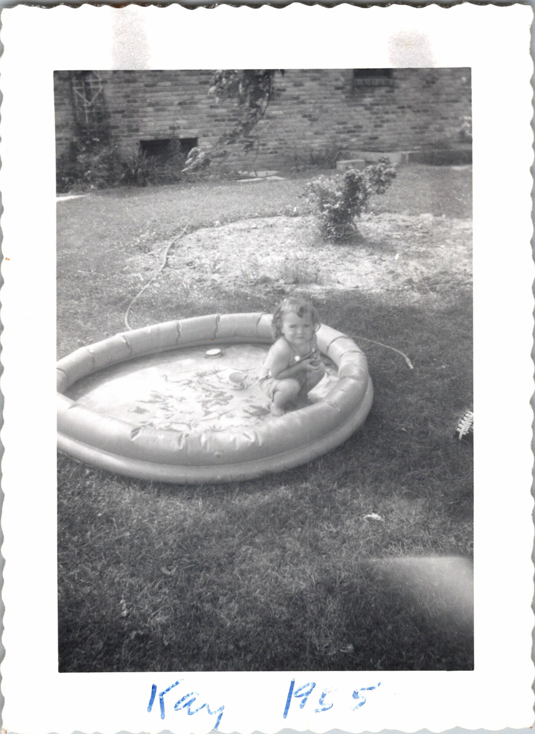 Toddler sitting in a round inflatable pool on the lawn with a brick wall garden backdrop behind.