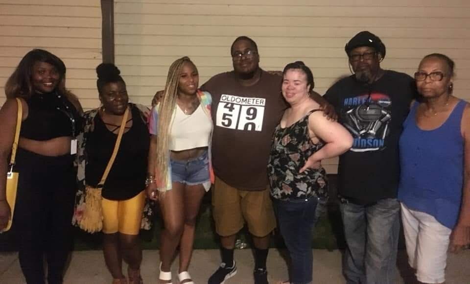 Group of eight people posing together outdoors at night, smiling for the camera, standing close with arms around each other in casual summer outfits