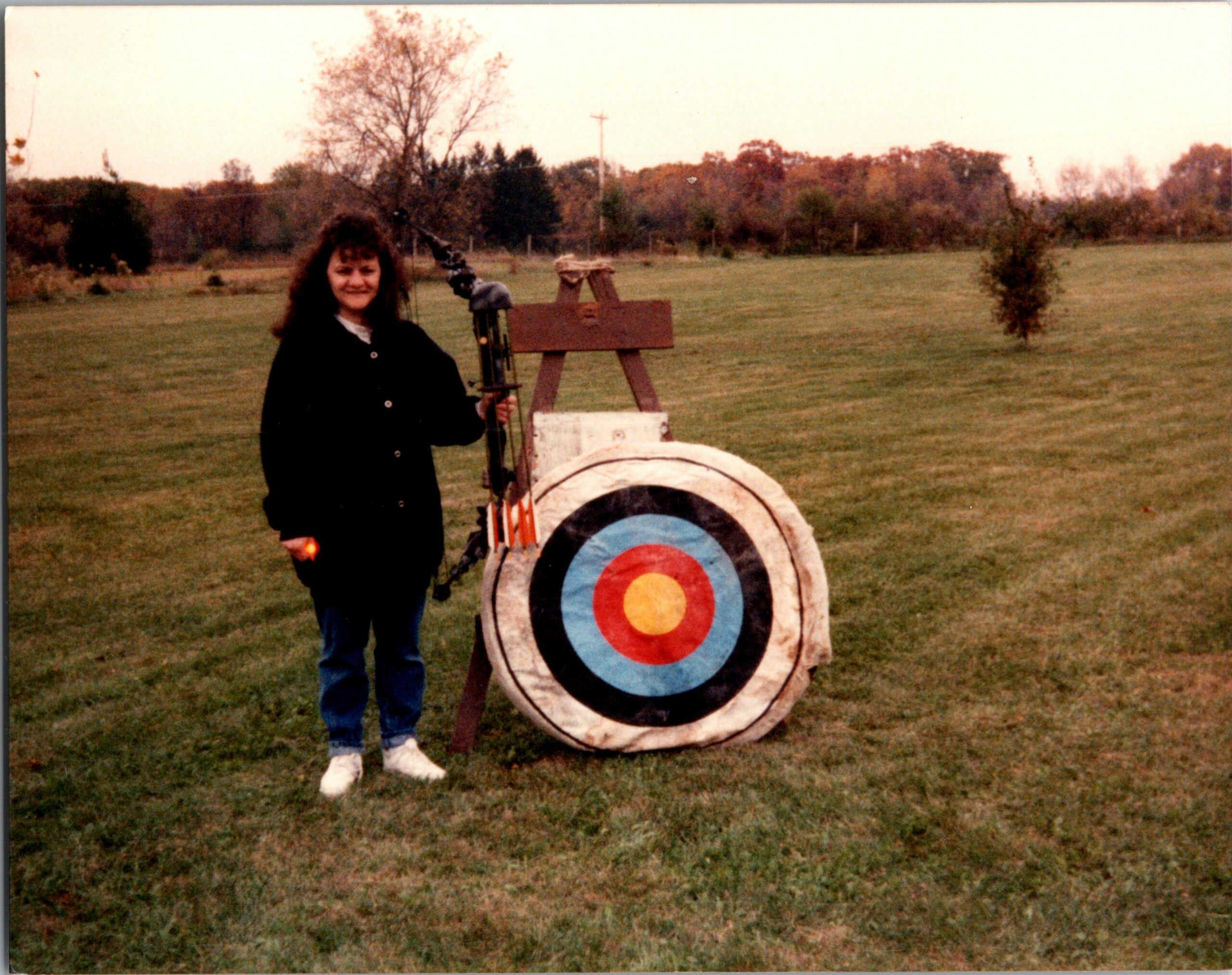 Person standing in a grassy field next to an archery target, holding a bow and smiling outdoors.