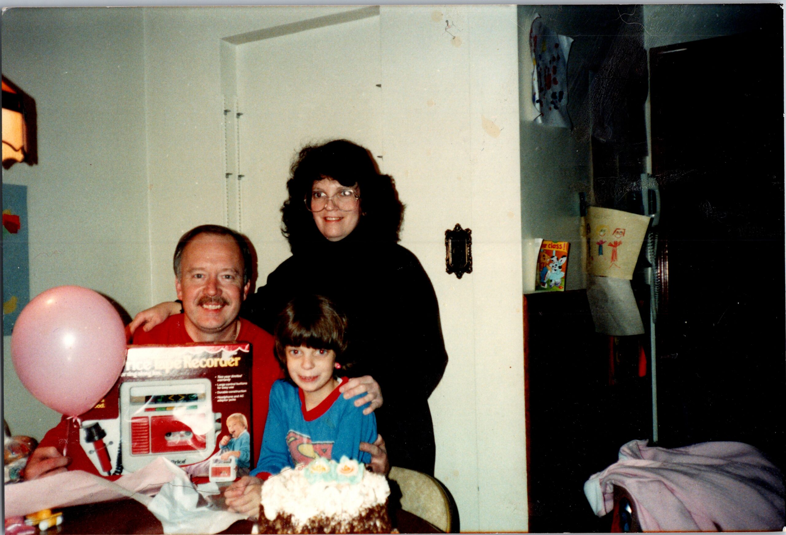 Family gathers around a birthday cake and gifts, posing for a photo with a man, a woman, and two children present in a home setting.