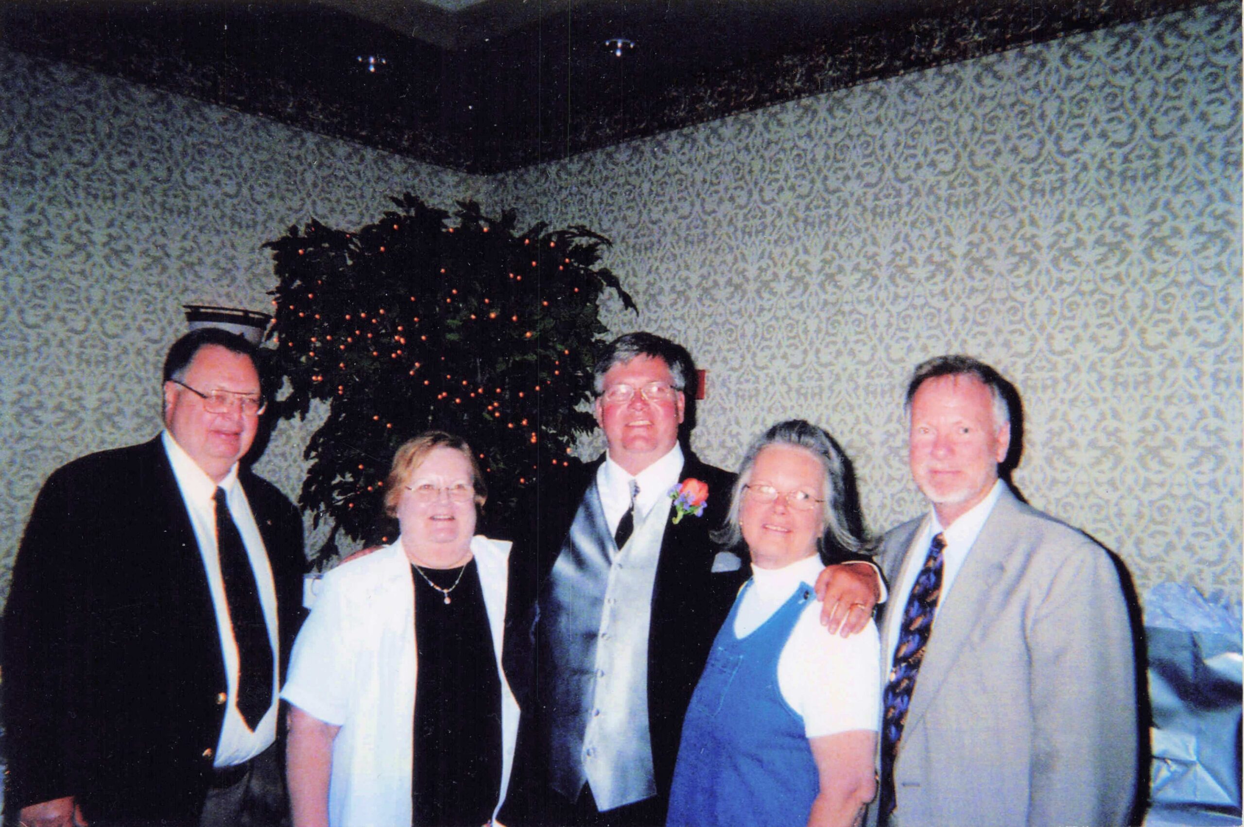 Group of six adults in formal attire posing together indoors, with a decorated Christmas tree in the background.