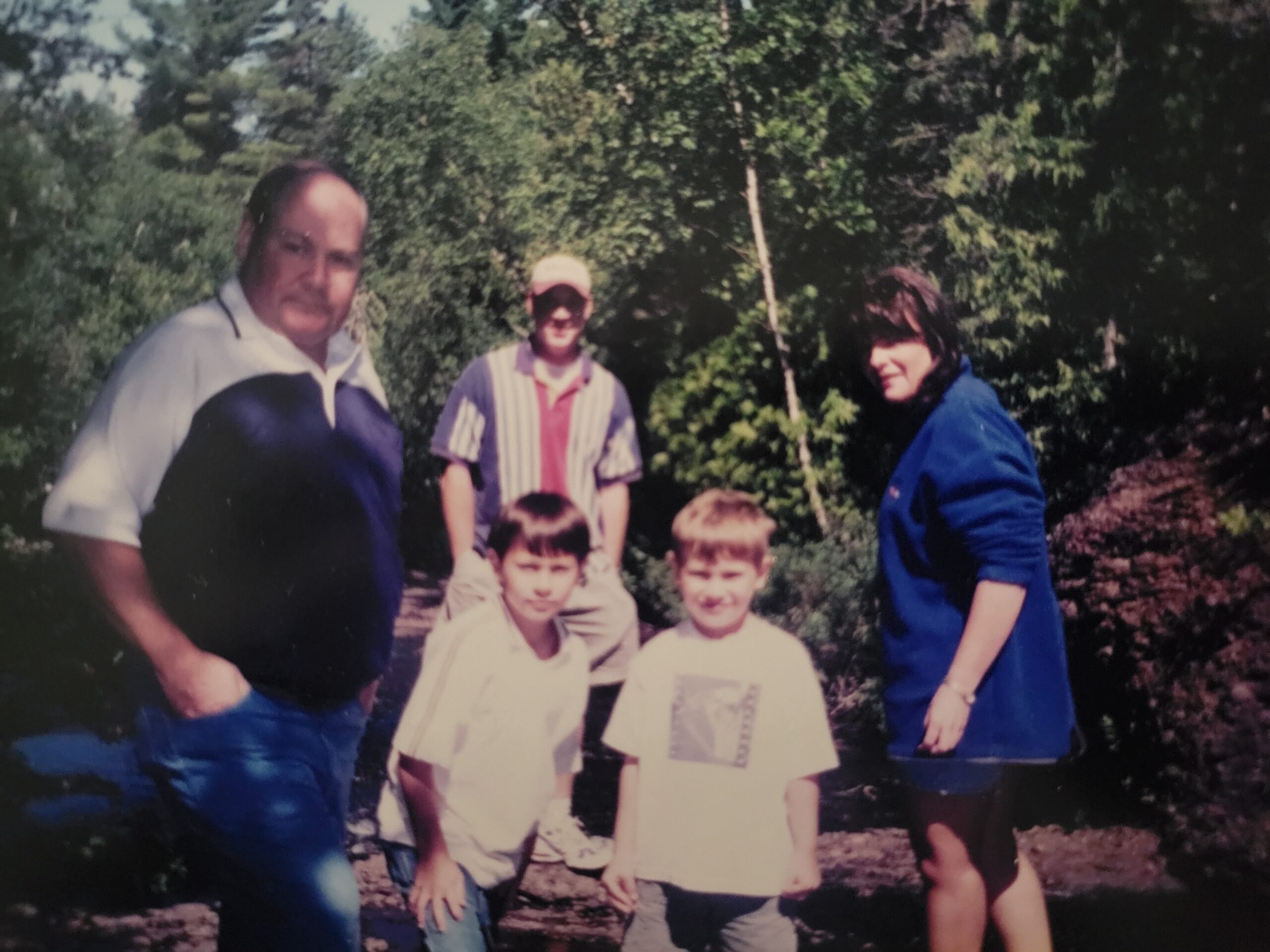 Group of five people outdoors in a wooded area; two adults on the sides and three children standing/two kneeling in front holding papers or posters.