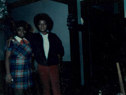 Two people posing together indoors in a dimly lit room; one wearing a plaid dress and the other in a dark jacket with red pants.