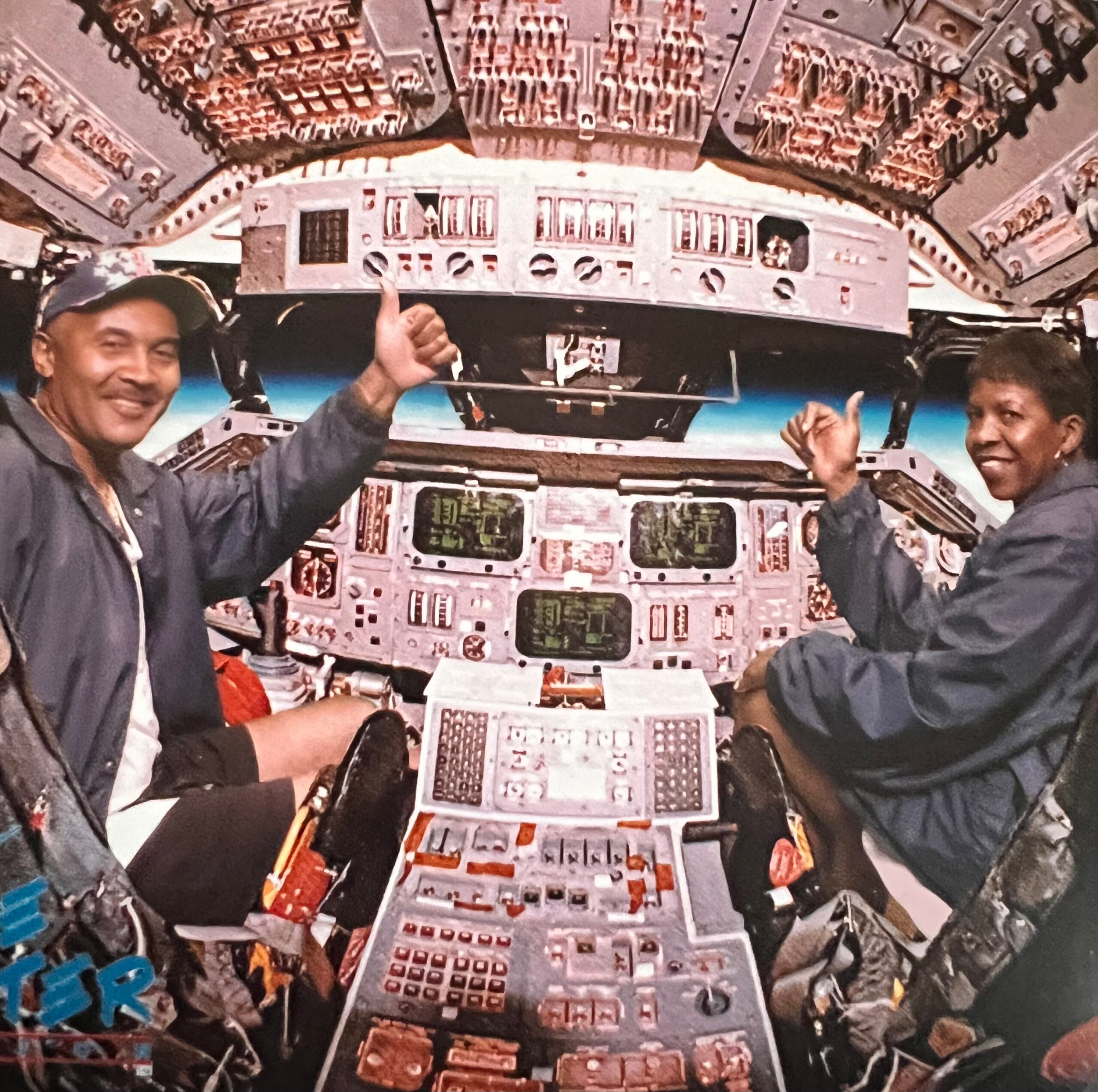 Two pilots smile and give thumbs up in a cockpit surrounded by avionics and gauges.