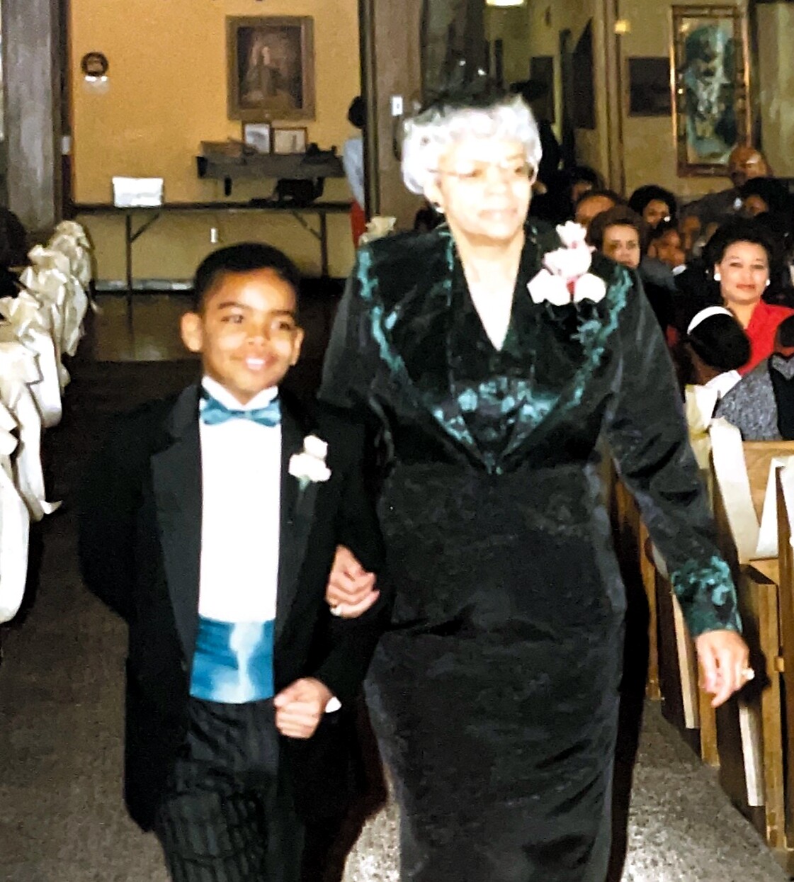 Young boy in a tuxedo walks arm-in-arm with an older woman in a dark teal dress down a church aisle; guests seated along the sides.