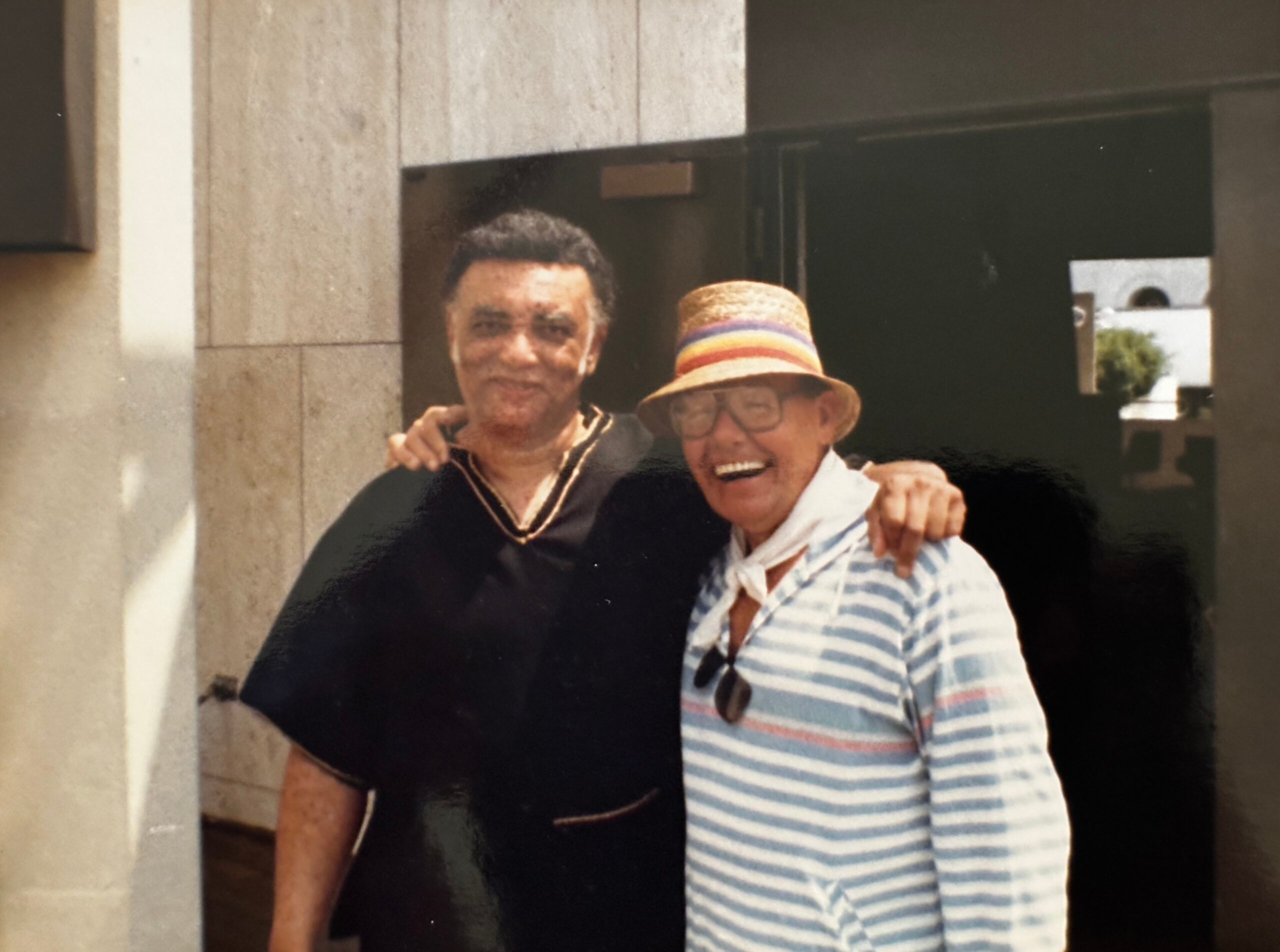 Two men posing together for a photo, one in a black shirt and the other in a striped shirt with a straw hat, in front of a doorway.