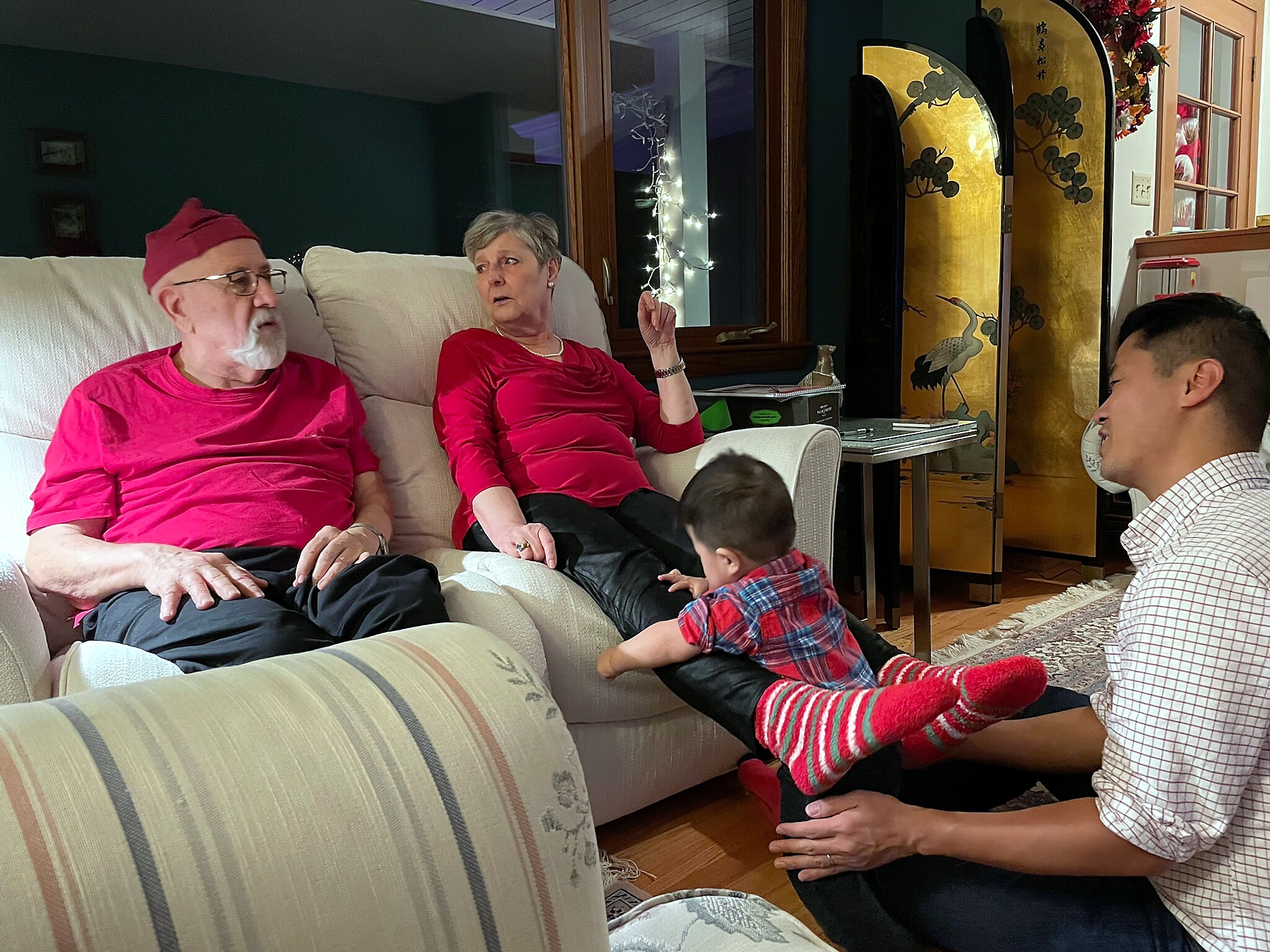Elderly couple in red shirts sits on a beige sofa in a cozy living room; a child sits on the floor nearby with festive lights in the background.