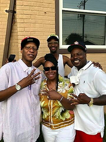 Four friends posing outside a brick building, smiling at the camera; two men wear caps, a woman in a yellow floral top sits center, with a man in a white tank behind her.