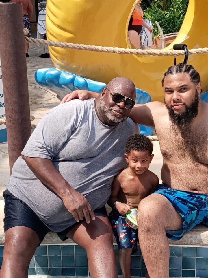 Three people sit together by a pool at a water park: an older man in a gray shirt and sunglasses, a bearded man with cornrows, and a small boy in swim trunks, smiling at the camera.