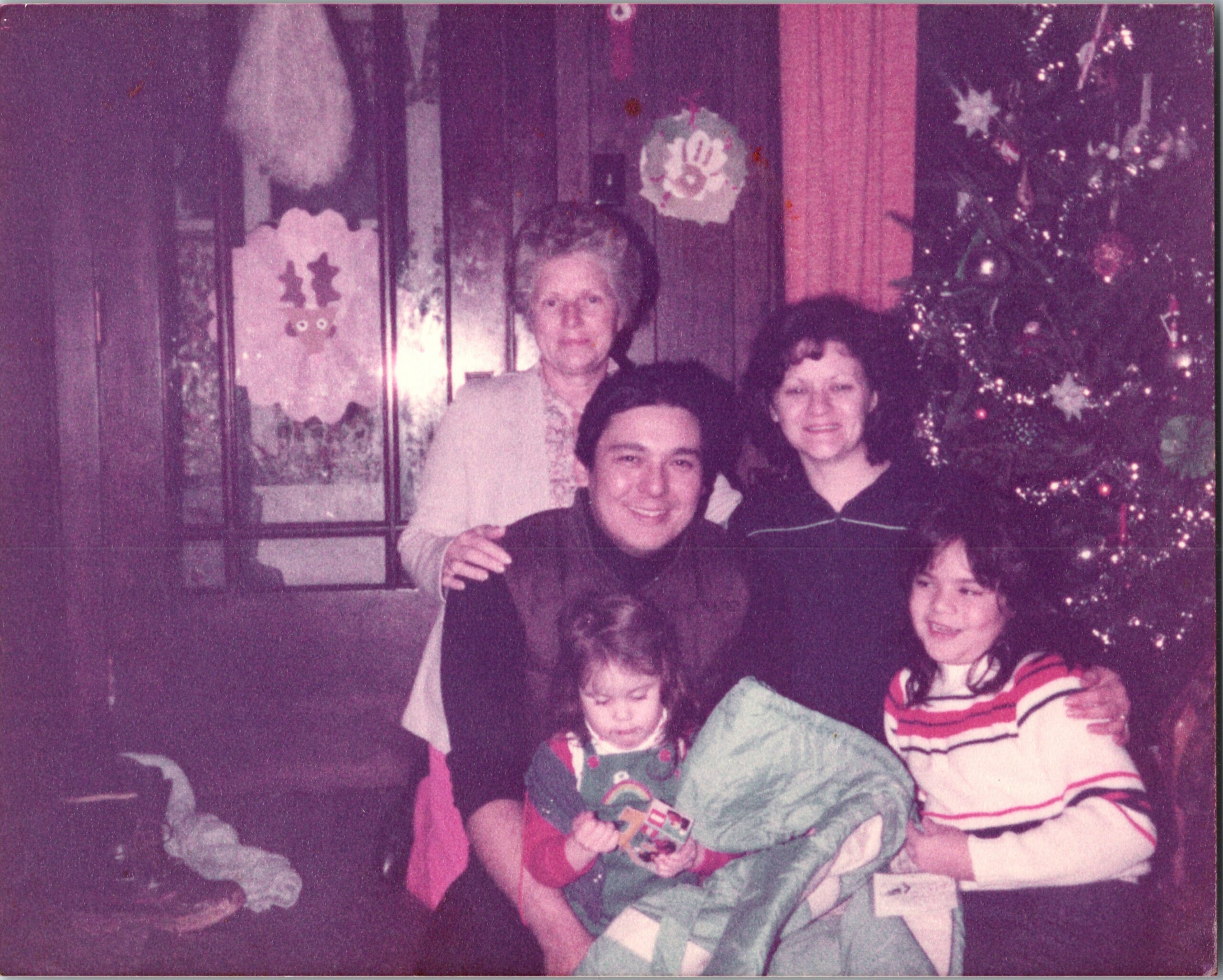 Family photo in a living room during Christmas; adults in back, children in front, with a lit Christmas tree on the right.
