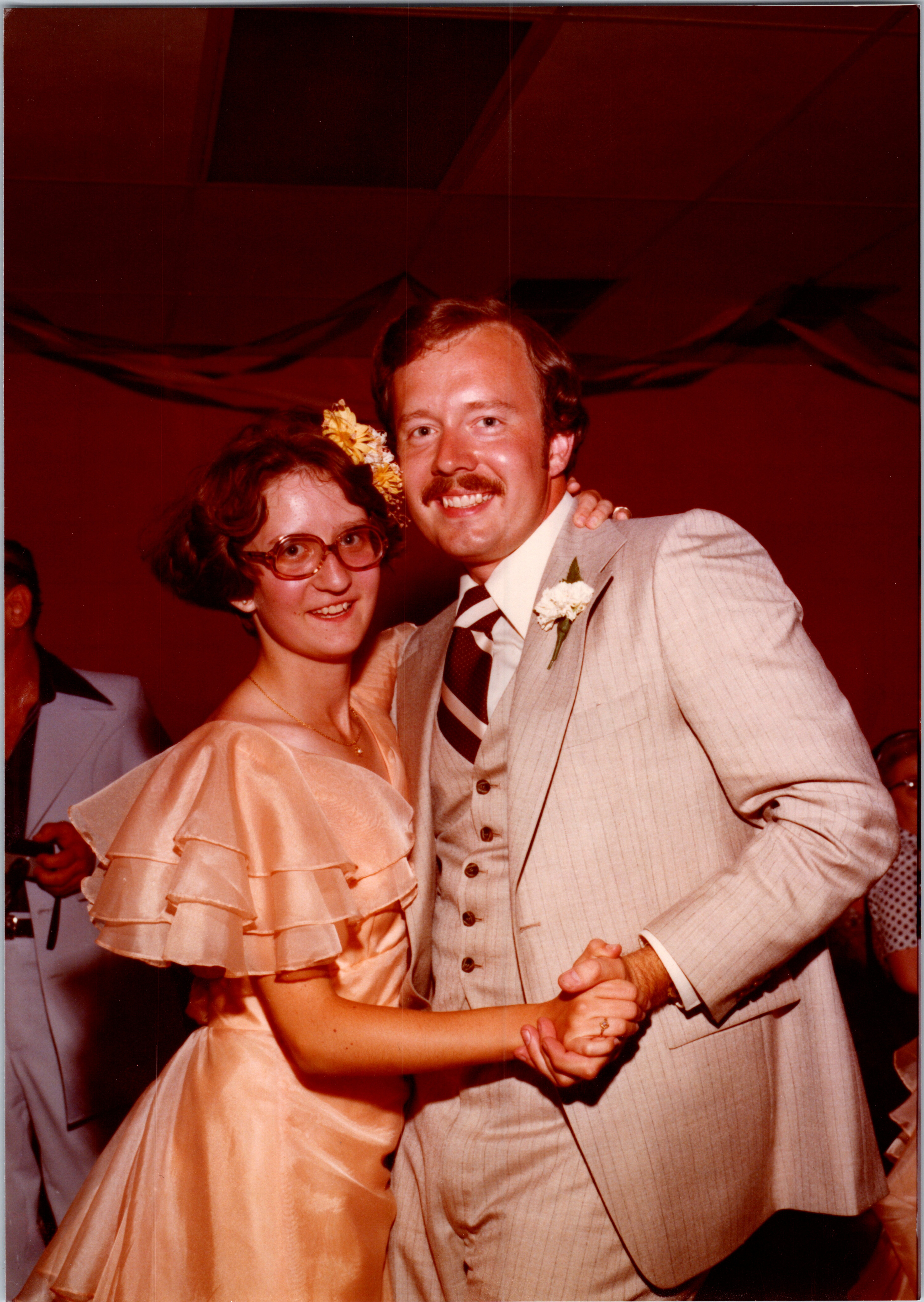 Smiling couple dancing at a wedding: woman in a peach dress with a flower in her hair, man in a light suit with a boutonniere.