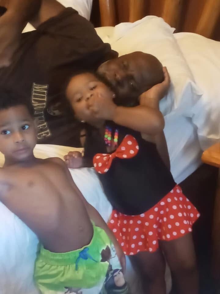 Family moment on a bed: a man lies with a young girl as he kisses her head, while a shirtless boy stands nearby.