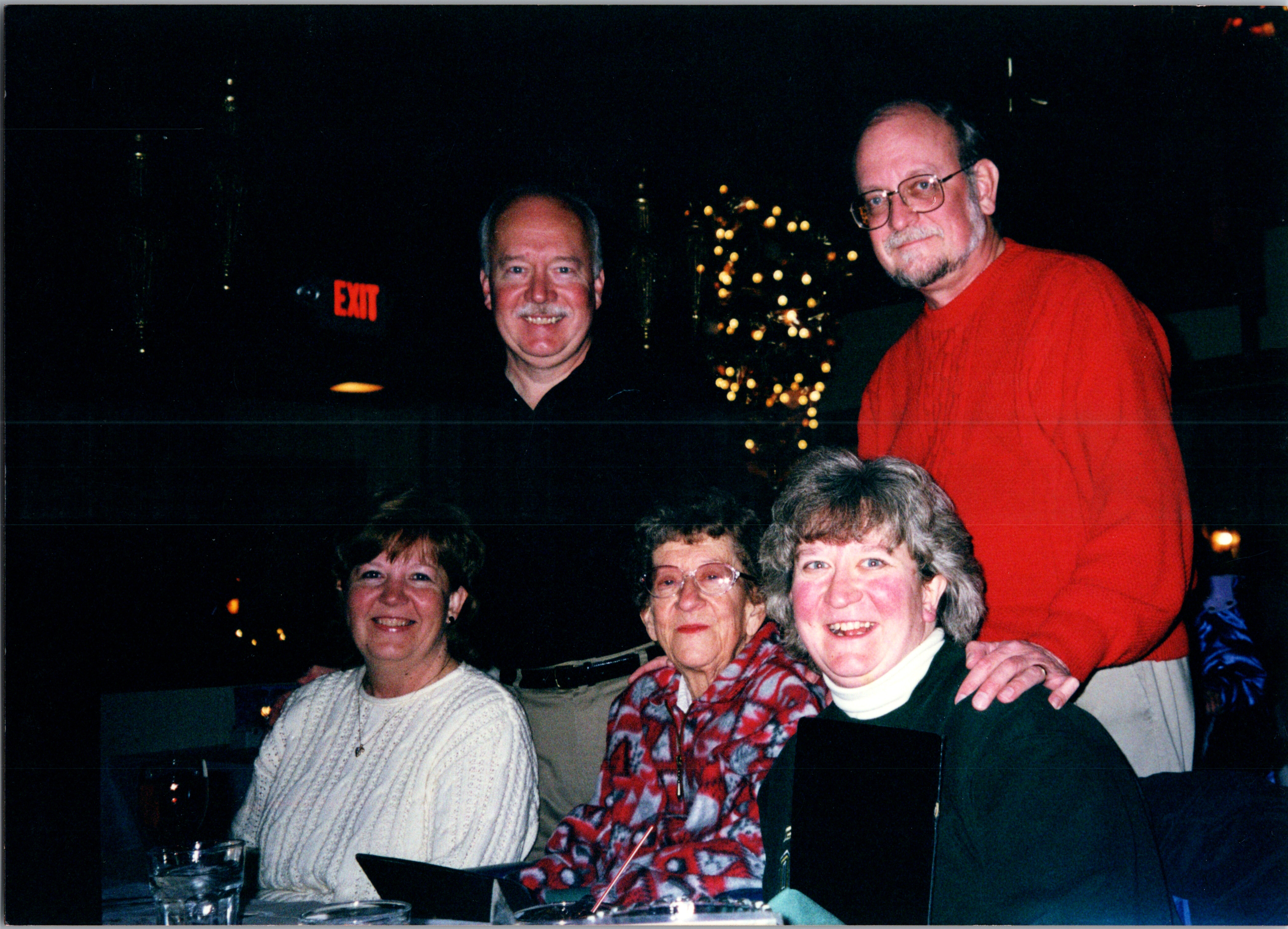 Group of smiling seniors at a holiday gathering around a table, Christmas tree lights in the background.