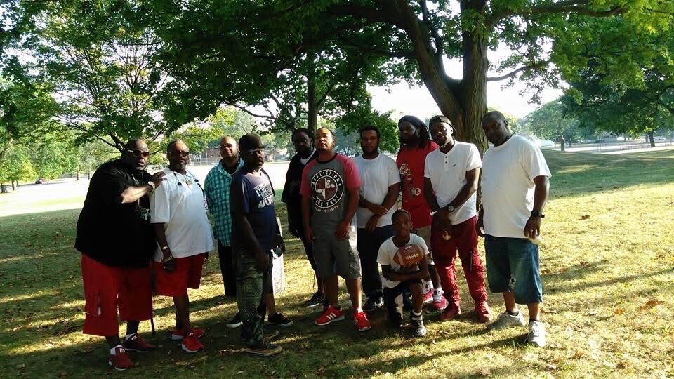 Group of men and a boy posing together in a sunny park under leafy trees.