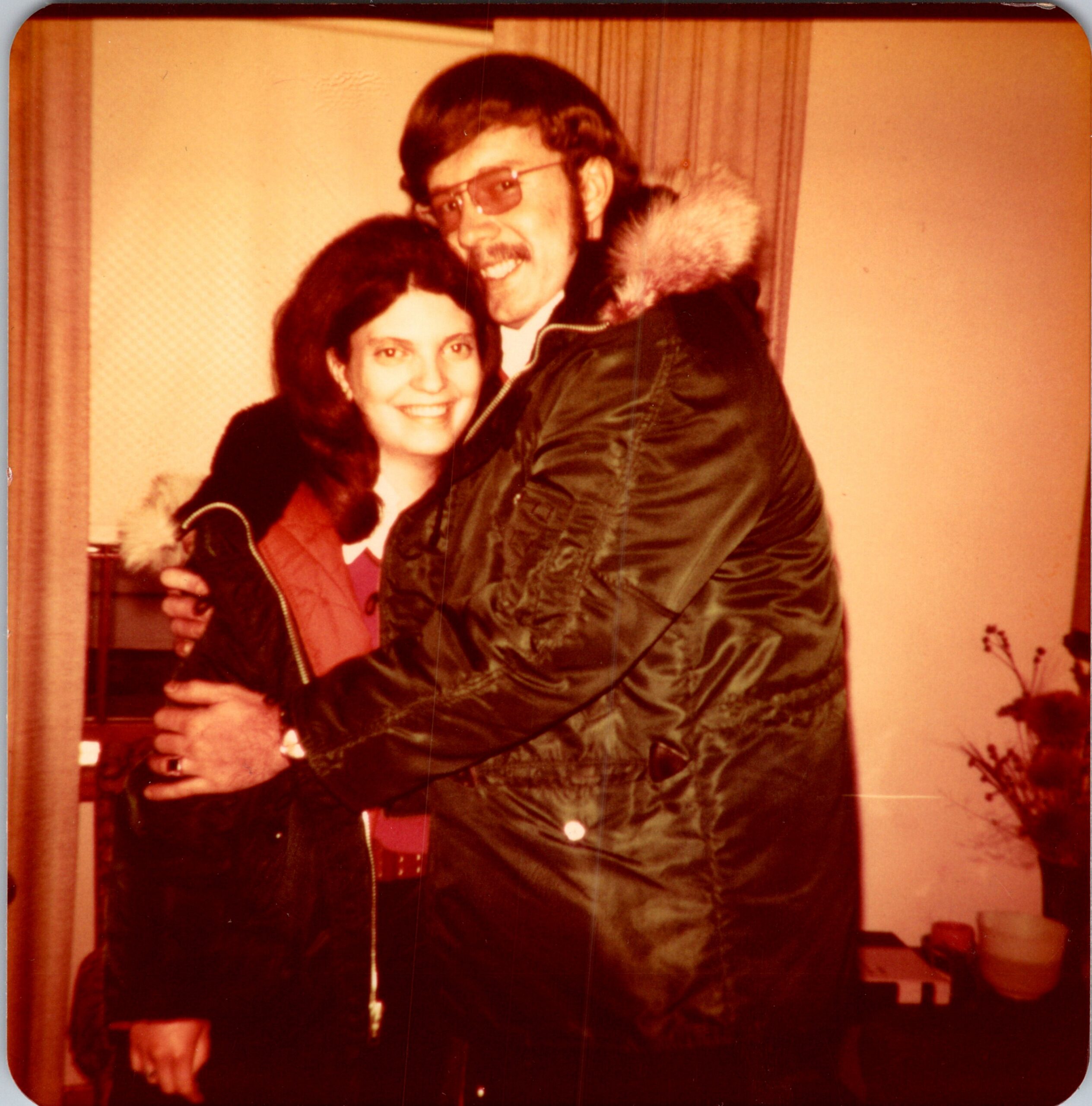 A smiling couple hugs each other indoors, with the woman in a red coat and the man in a dark jacket with a fur-lined hood.