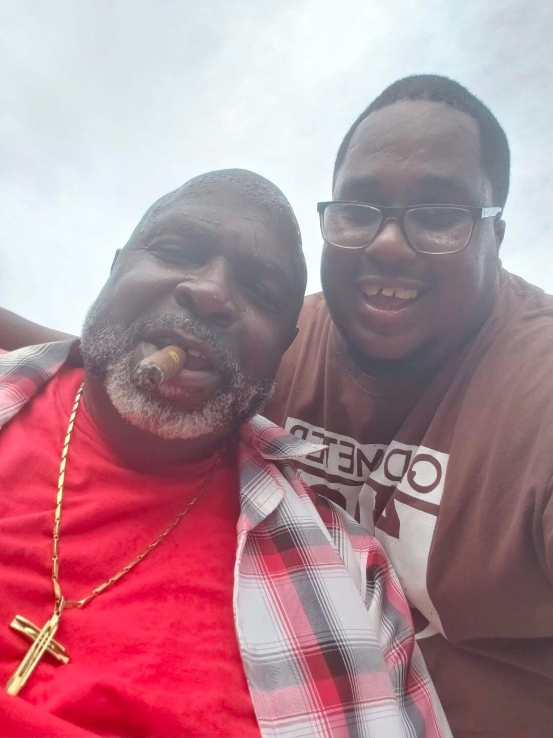 Two men taking a selfie outdoors; one has a cigar, a gold cross necklace, a red shirt and plaid jacket on.