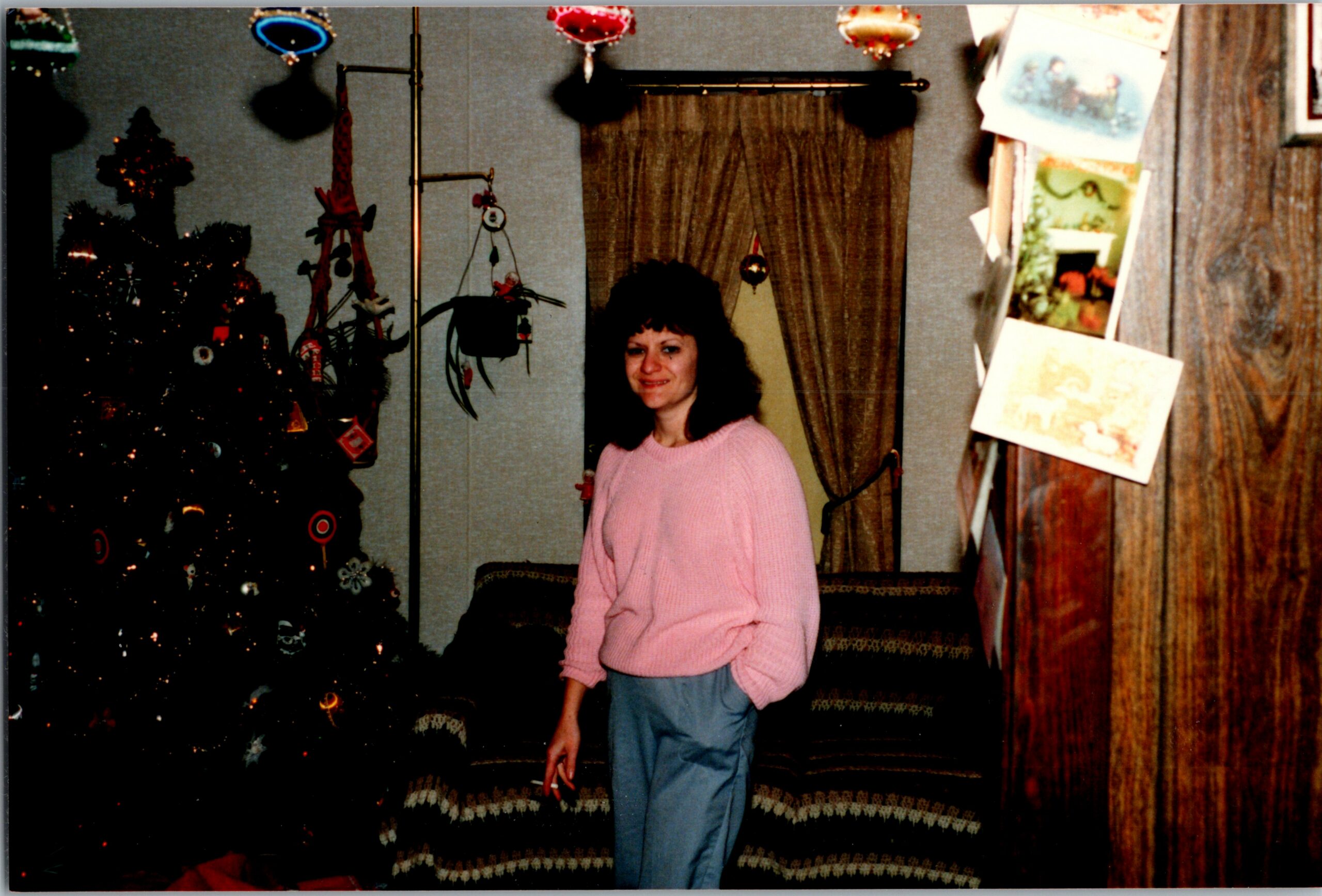 Woman in a pink sweater stands in a living room with a decorated Christmas tree to her left and cards hanging on a wooden wall to the right, smiling at the camera.