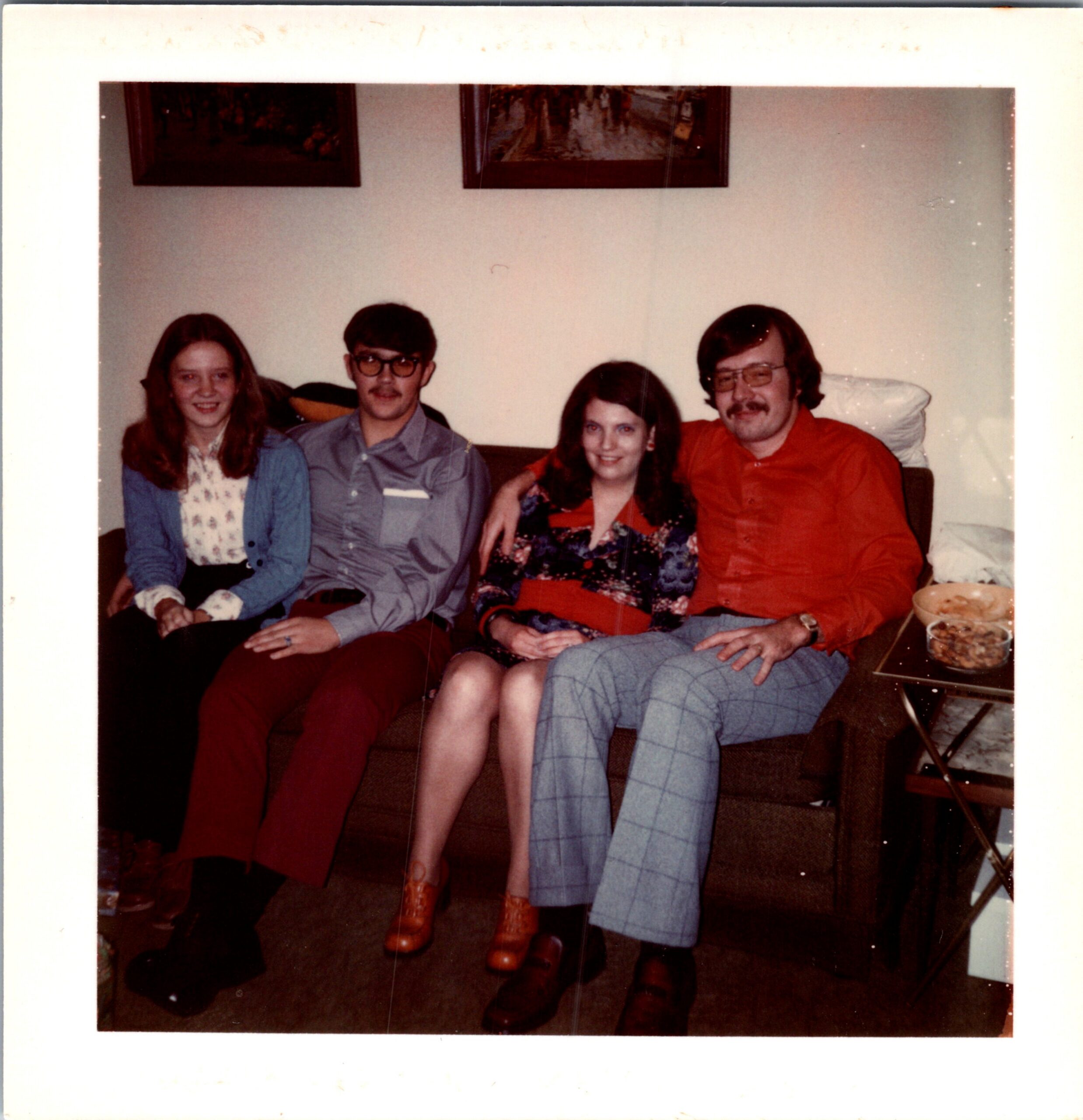 Four friends sit closely on a couch in a living room, smiling at the camera; a small table with bowls sits to the right.