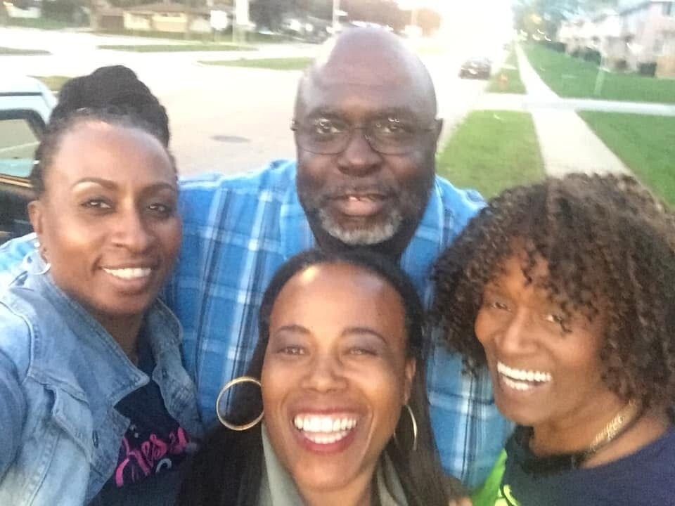 Group of four friends posing for a selfie outdoors on a sunny street, all smiling at the camera.