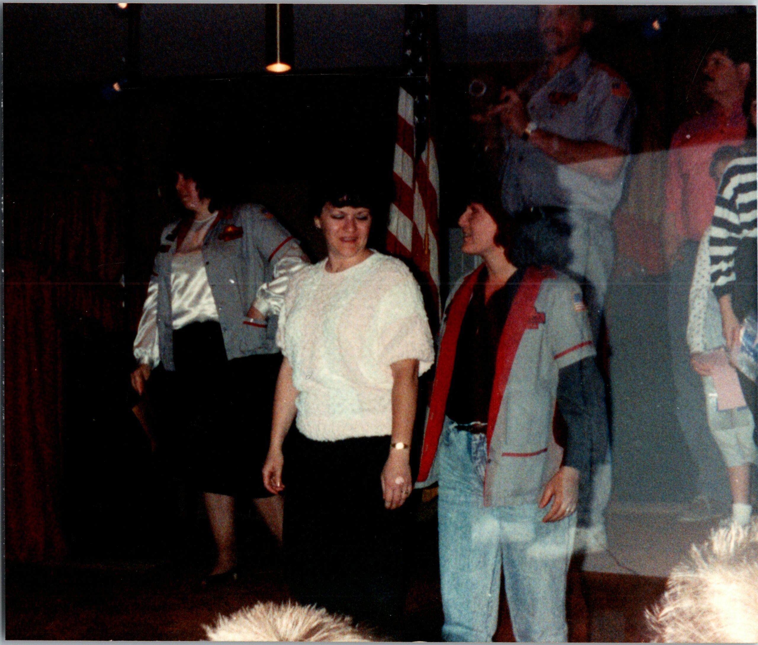 People dancing and chatting in a dimly lit room, with an American flag visible in the background and a woman in a white top center smiling.