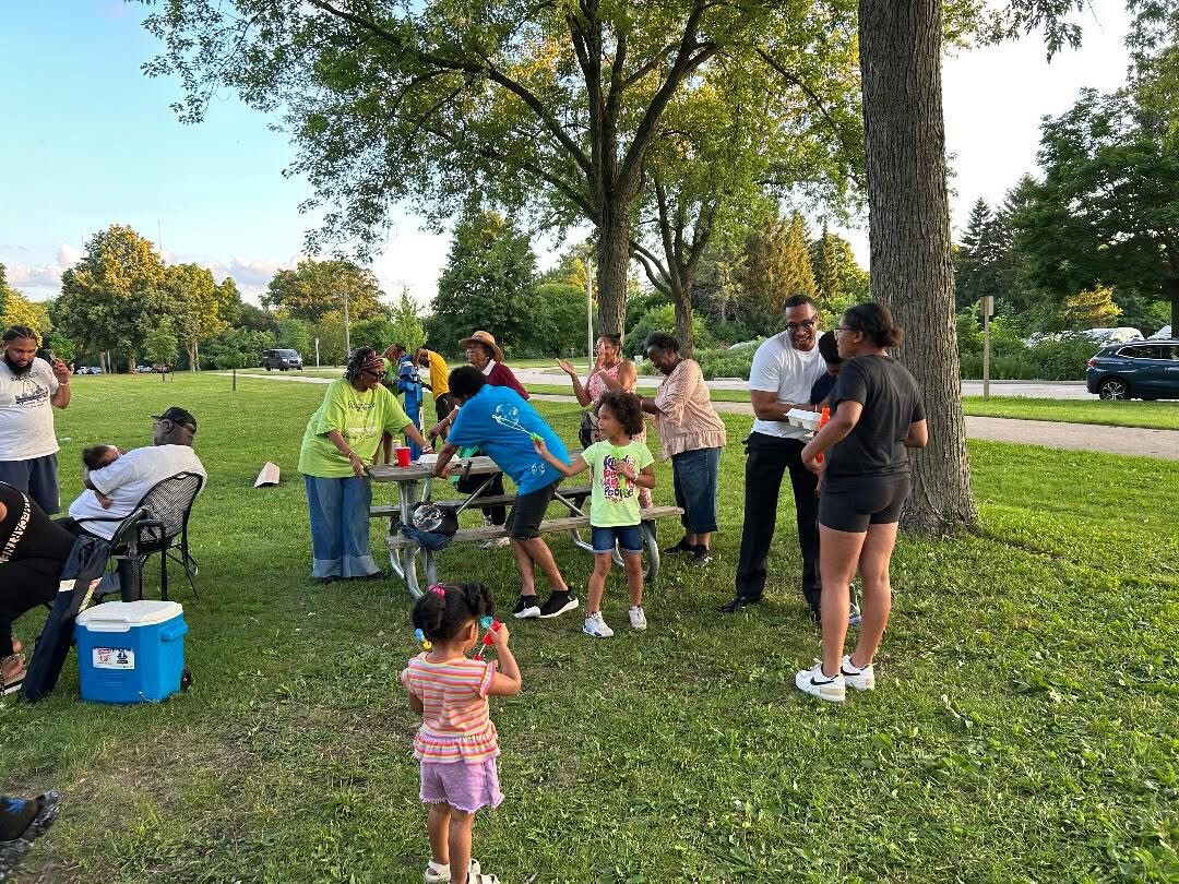 Group of diverse people socializing at a park picnic under trees; children play nearby by a table and cooler.