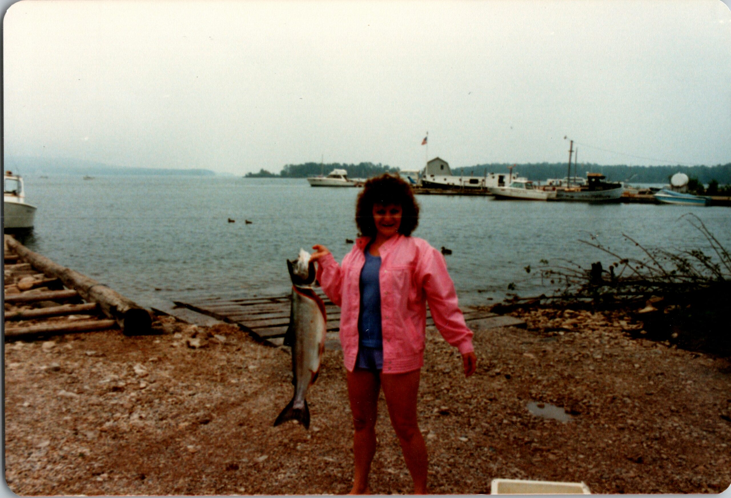 Woman in a pink jacket standing on a rocky shore, holding a large fish with boats in a calm bay behind her.