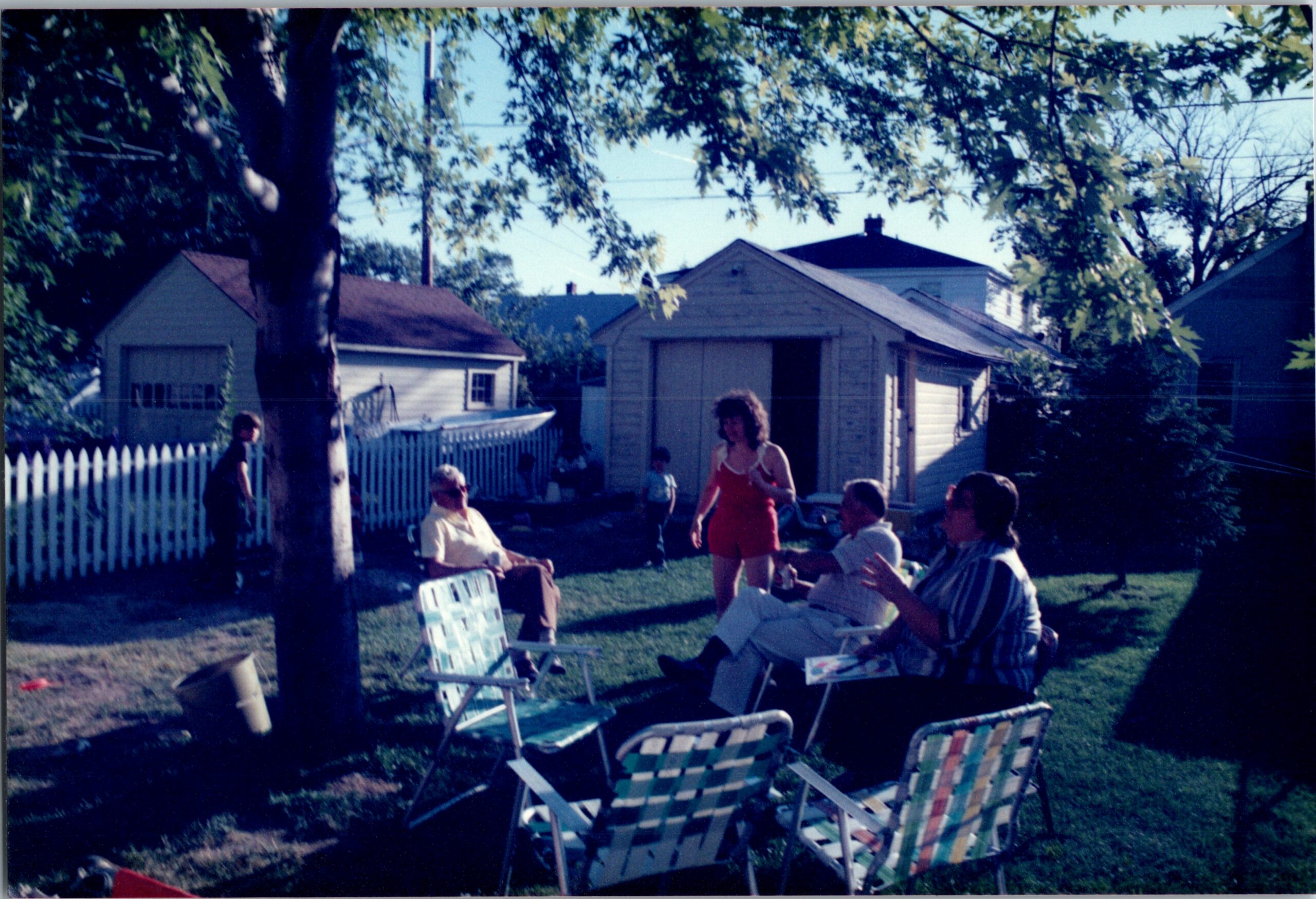 Group of people relaxing in a sunny backyard with folding lawn chairs and a white picket fence nearby. Behind them are small houses and a shed.