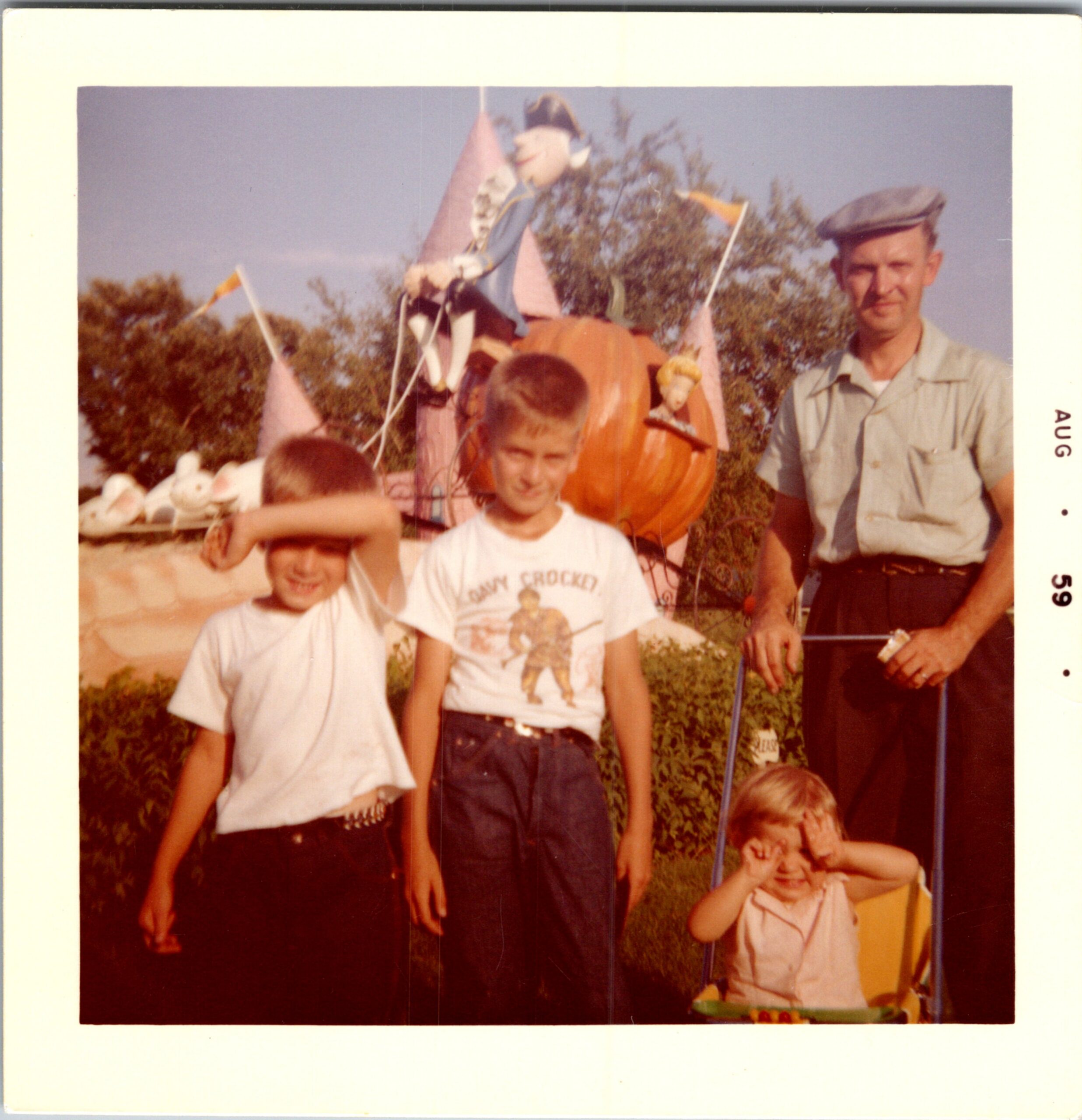 Group of kids at a parade posing beside a bright orange pumpkin float with pink sails; a man in a light shirt and cap stands to the right.
