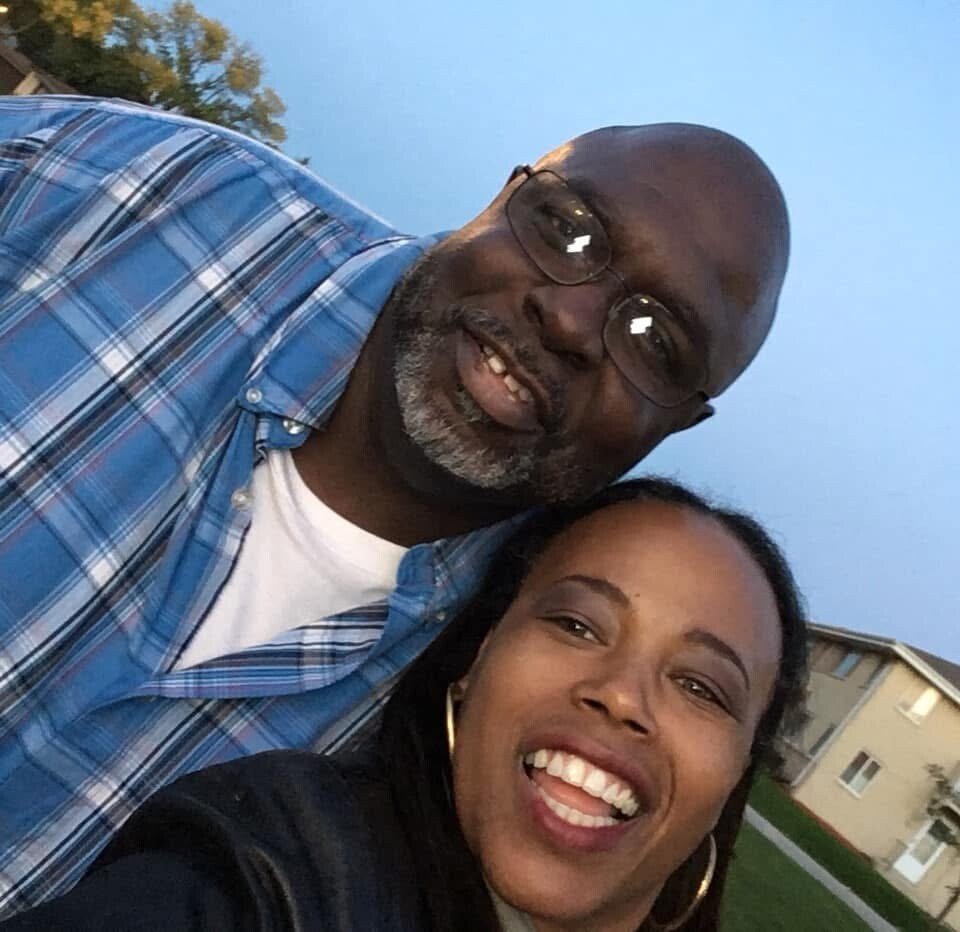 Smiling couple taking a selfie outdoors in a suburban neighborhood; man wears glasses and a blue plaid shirt, woman wears hoop earrings.