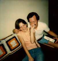 Two friends on a couch laughing together, a crocheted blanket beside them.