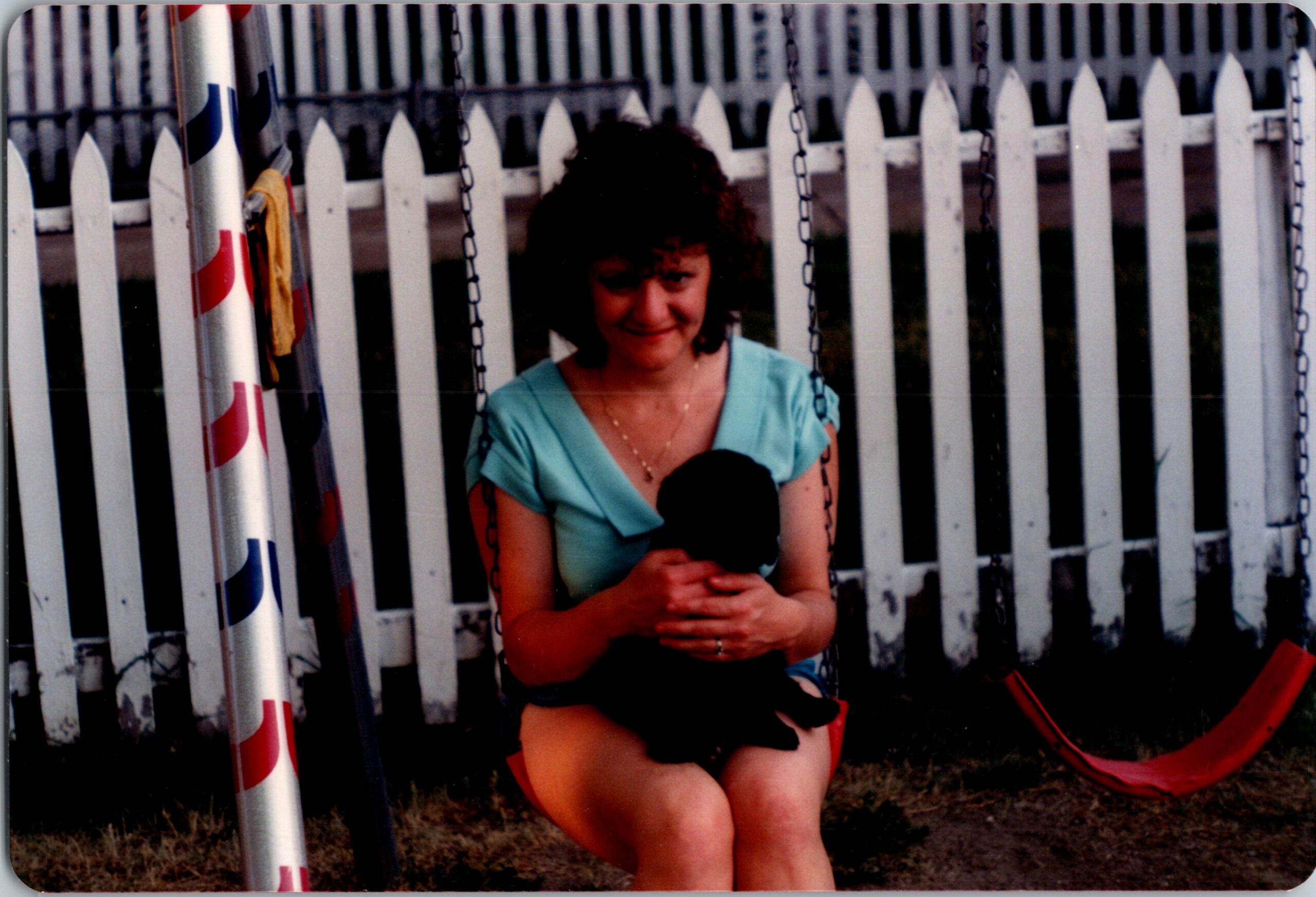 Woman in a teal top sits on a swing, holding a small black dog, with a white picket fence in the background.