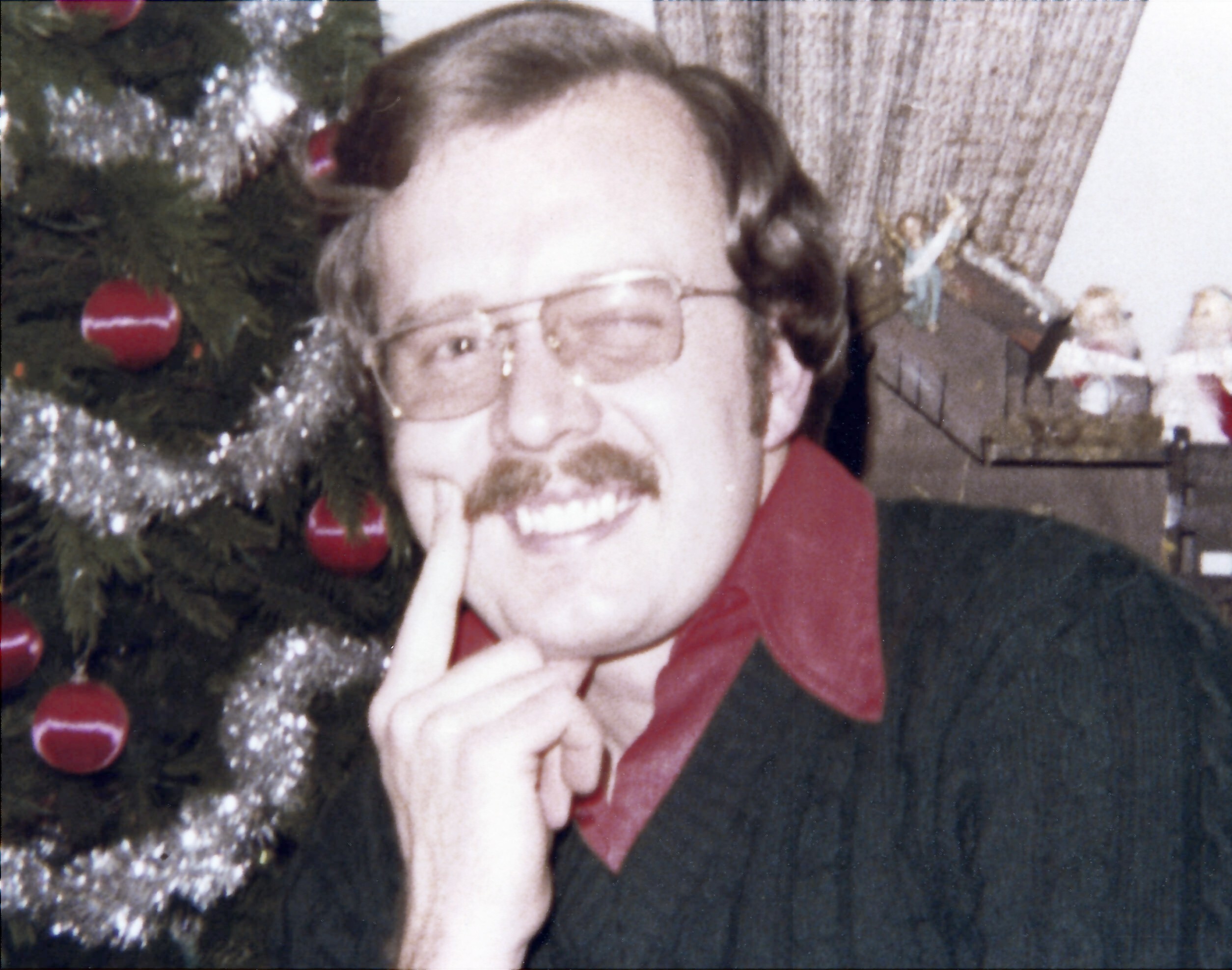 Close-up of a man with glasses smiling beside a decorated Christmas tree with red baubles.