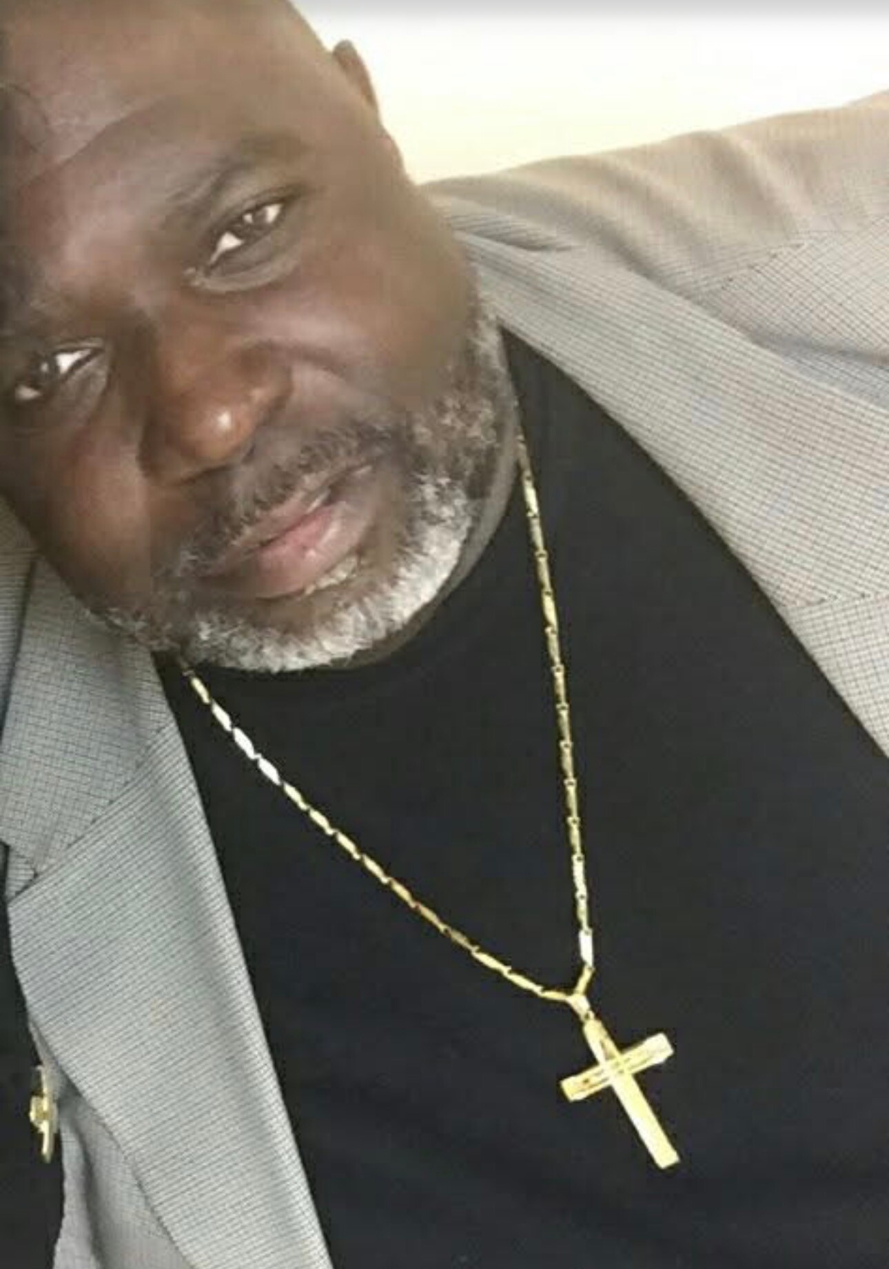 Close-up of a man wearing a light gray blazer over a black shirt, with a gold cross necklace visible at chest level.