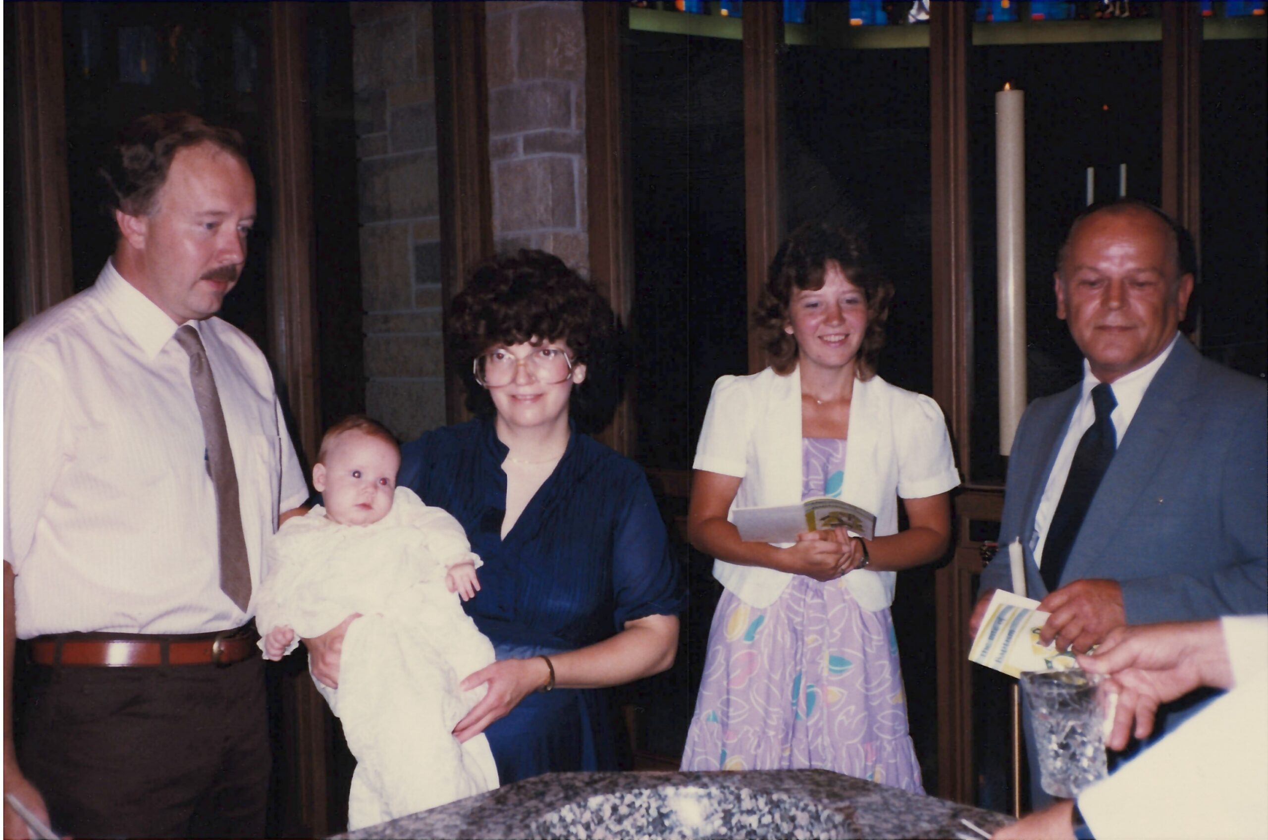 Family gathers for a church ceremony around a baptismal font, with a baby held by a man and others smiling nearby.