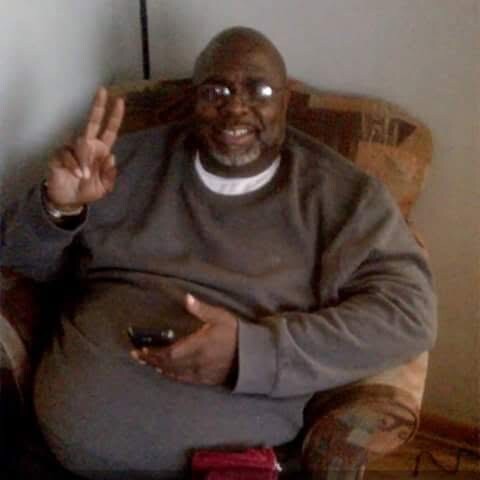 Man sitting in a brown armchair, smiling and flashing a peace sign with his right hand, holding a phone in his lap.