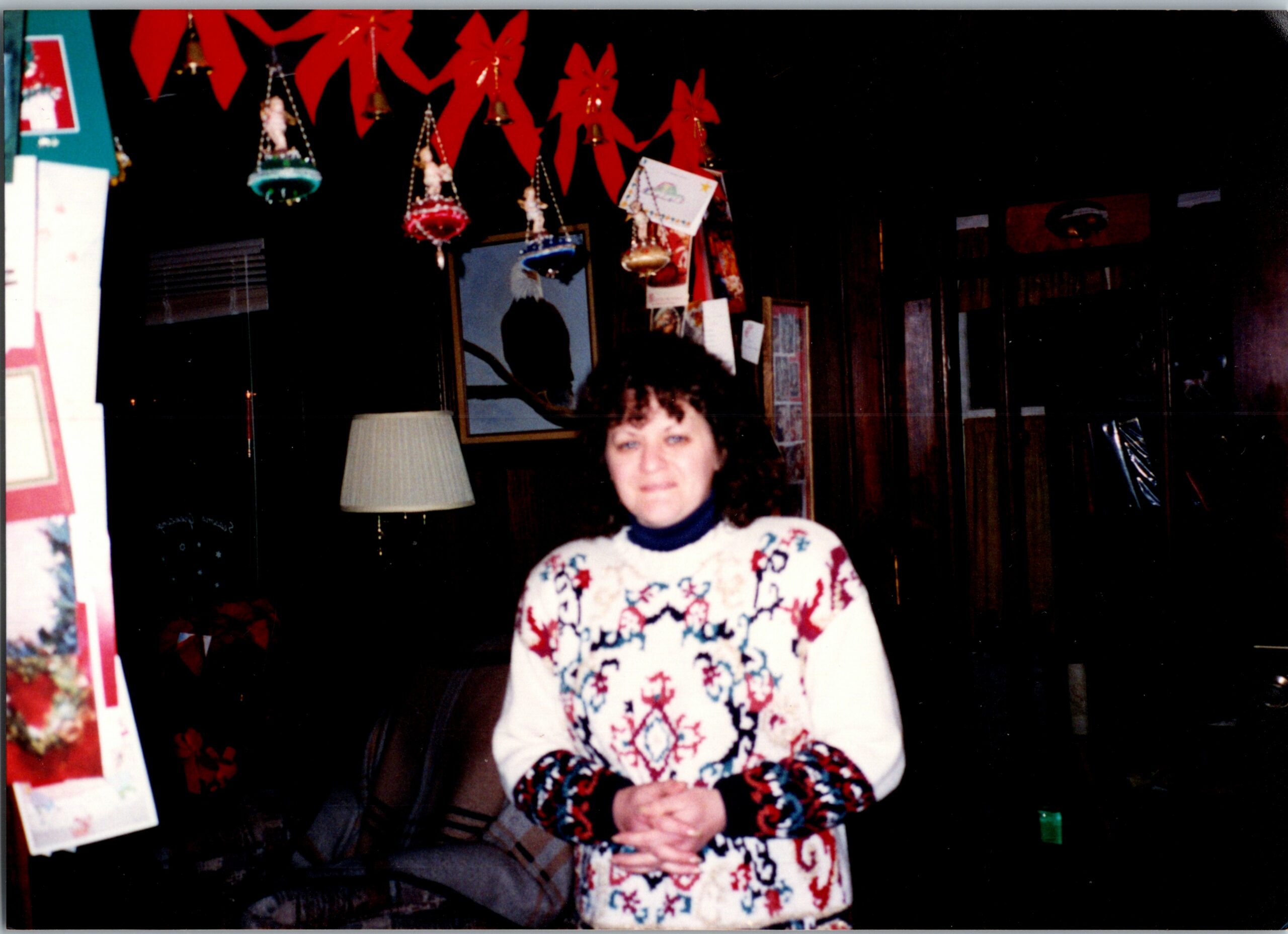 Woman in a white patterned Christmas sweater standing indoors with festive red ribbons and ornaments overhead.”, “Festive scene with a woman indoors, red decorations and hanging ornaments above her.”, “Smiling woman wearing a holiday sweater, surrounded by Christmas decor and a lamp.”, “Indoor holiday setting: woman in sweater, red ribbons and ornaments hanging from the ceiling.”, “Cozy Christmas display behind a woman in a patterned sweater, with framed art and decorations.”