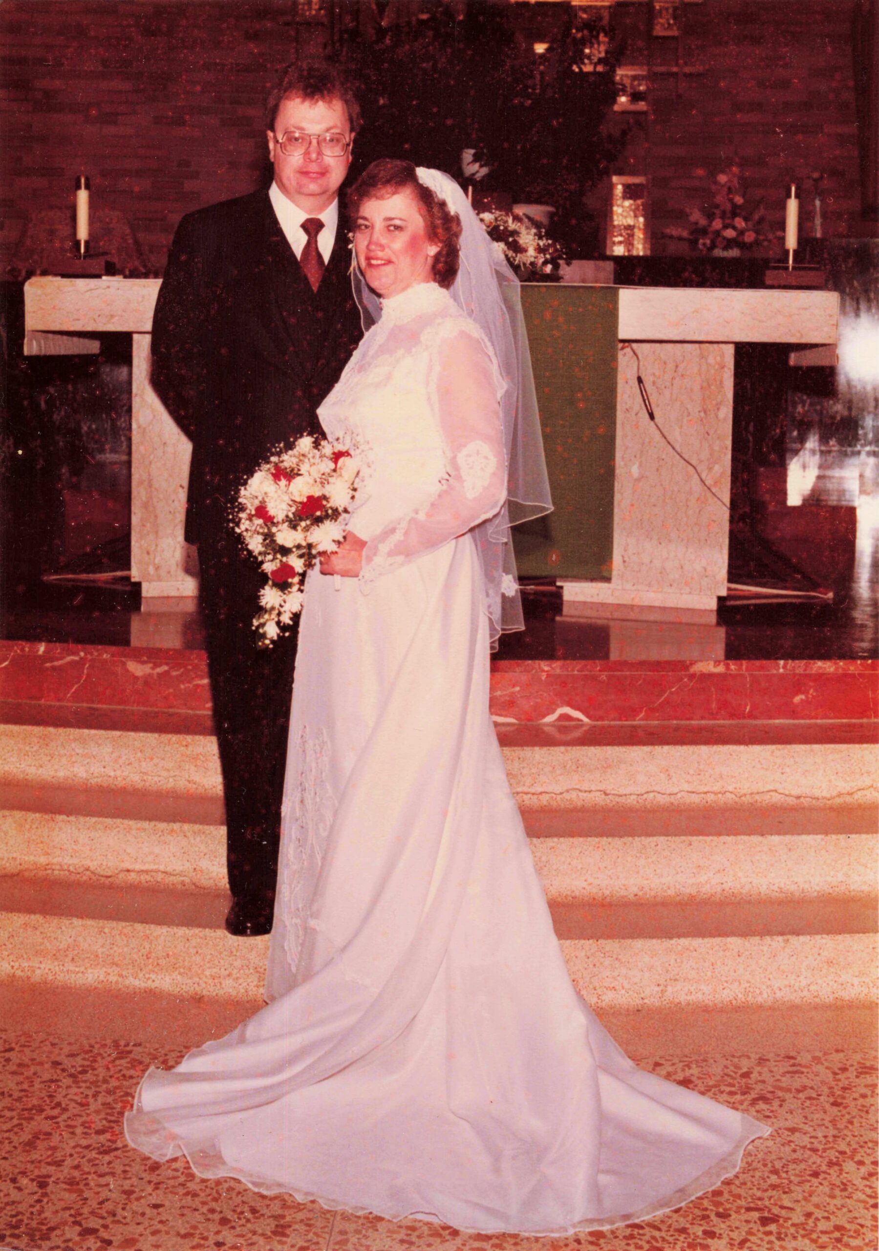 Bride and groom pose at the altar in a church, with the bride holding a bouquet and wearing a veil and white gown.