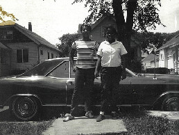 Black-and-white photo of two young men standing on a sidewalk in front of a vintage car, with a tall stack of books between them.