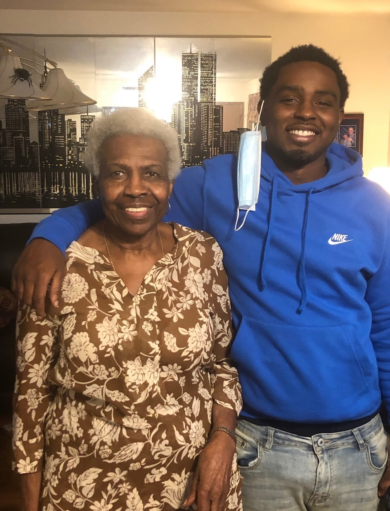 Smiling elderly woman in a brown floral dress with a young man in a blue Nike hoodie; his mask hangs from one ear, indoors.