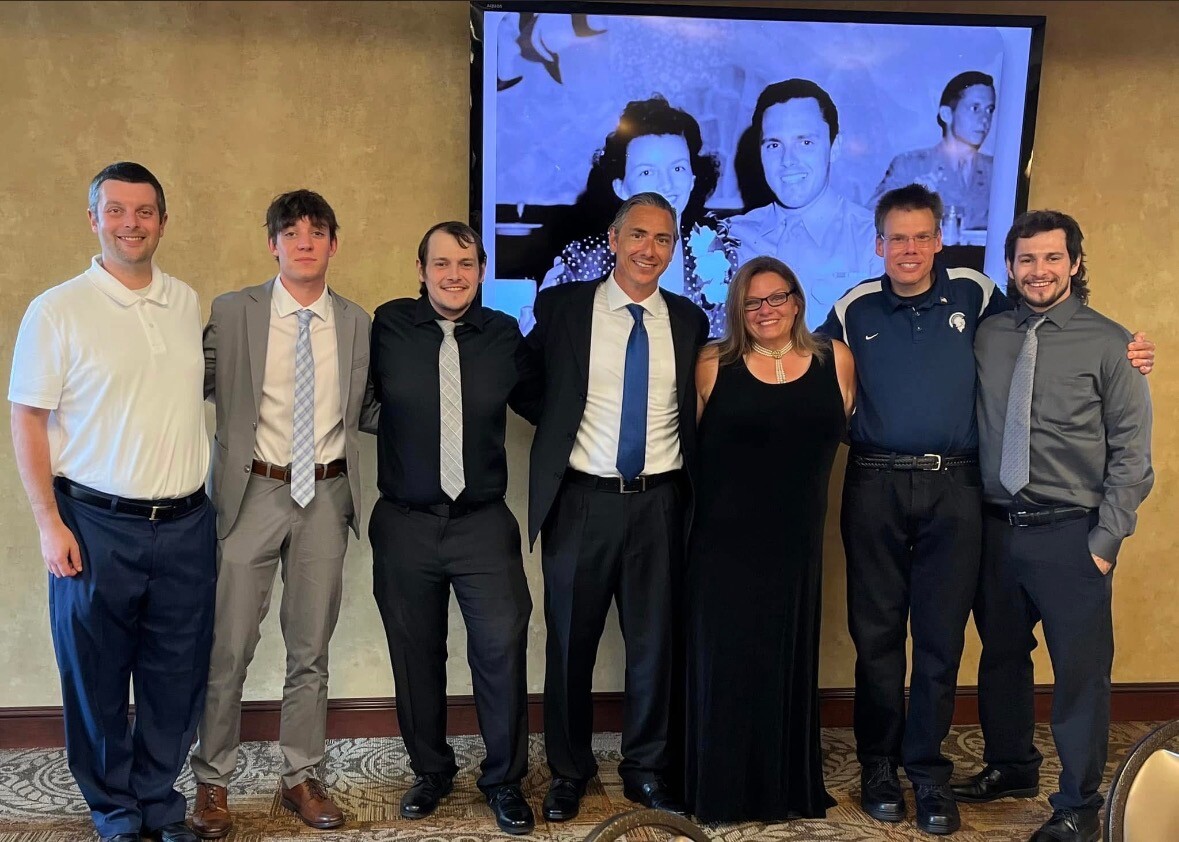 Seven adults posing for a group photo in a banquet hall, dressed in suits and dresses, in front of a large screen showing a black-and-white image behind them.
