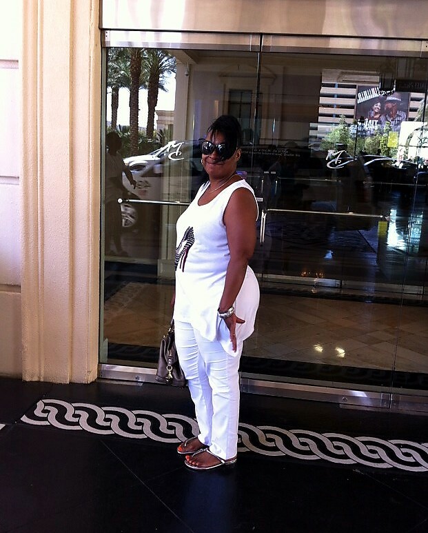 Woman in a white sleeveless top and white pants standing outside a glass storefront, wearing sunglasses and carrying a purse.