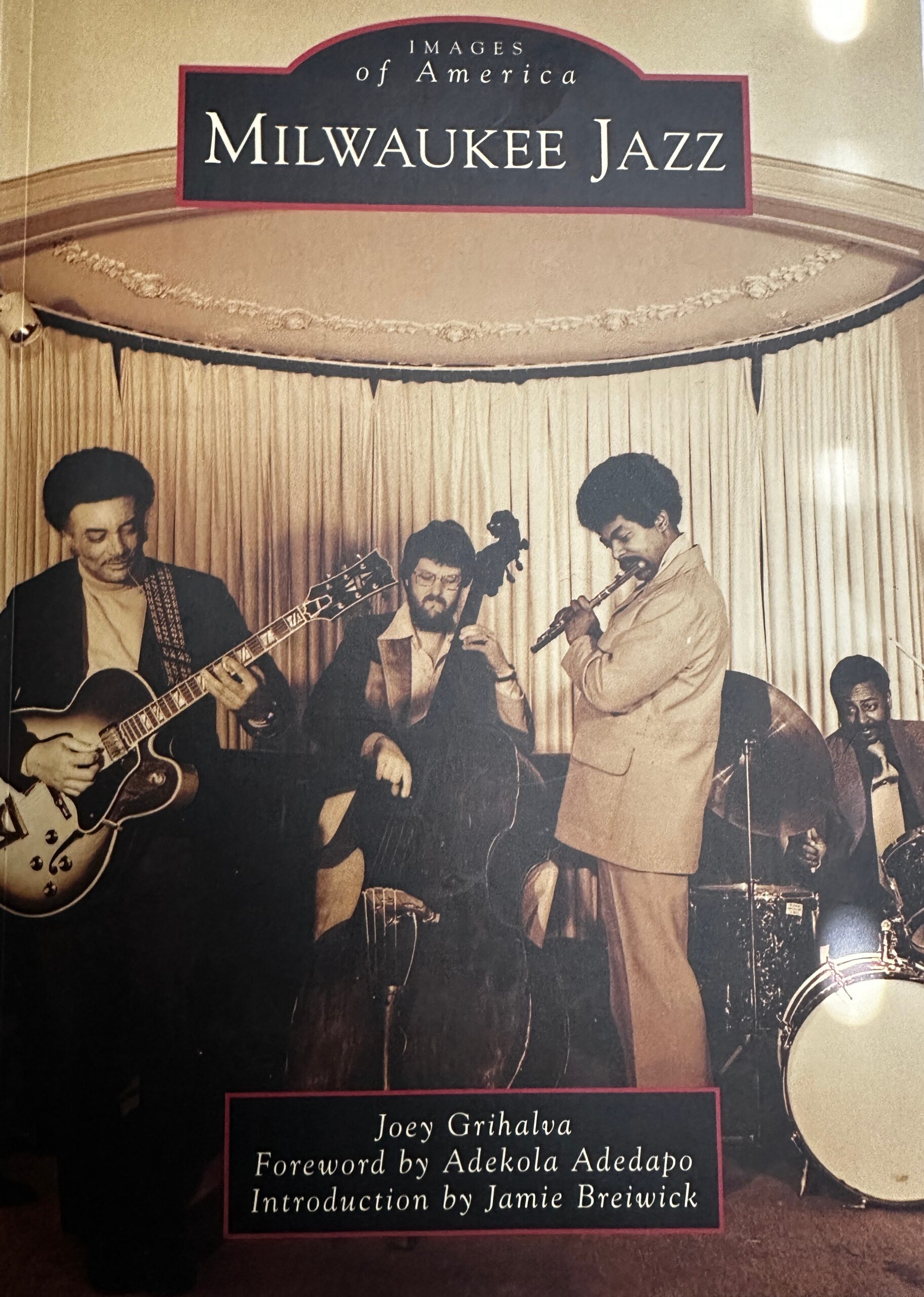 Three jazz musicians perform on a stage beneath a curved Milwaukee Jazz banner: guitarist on the left, singer in the center, reed player on the right.