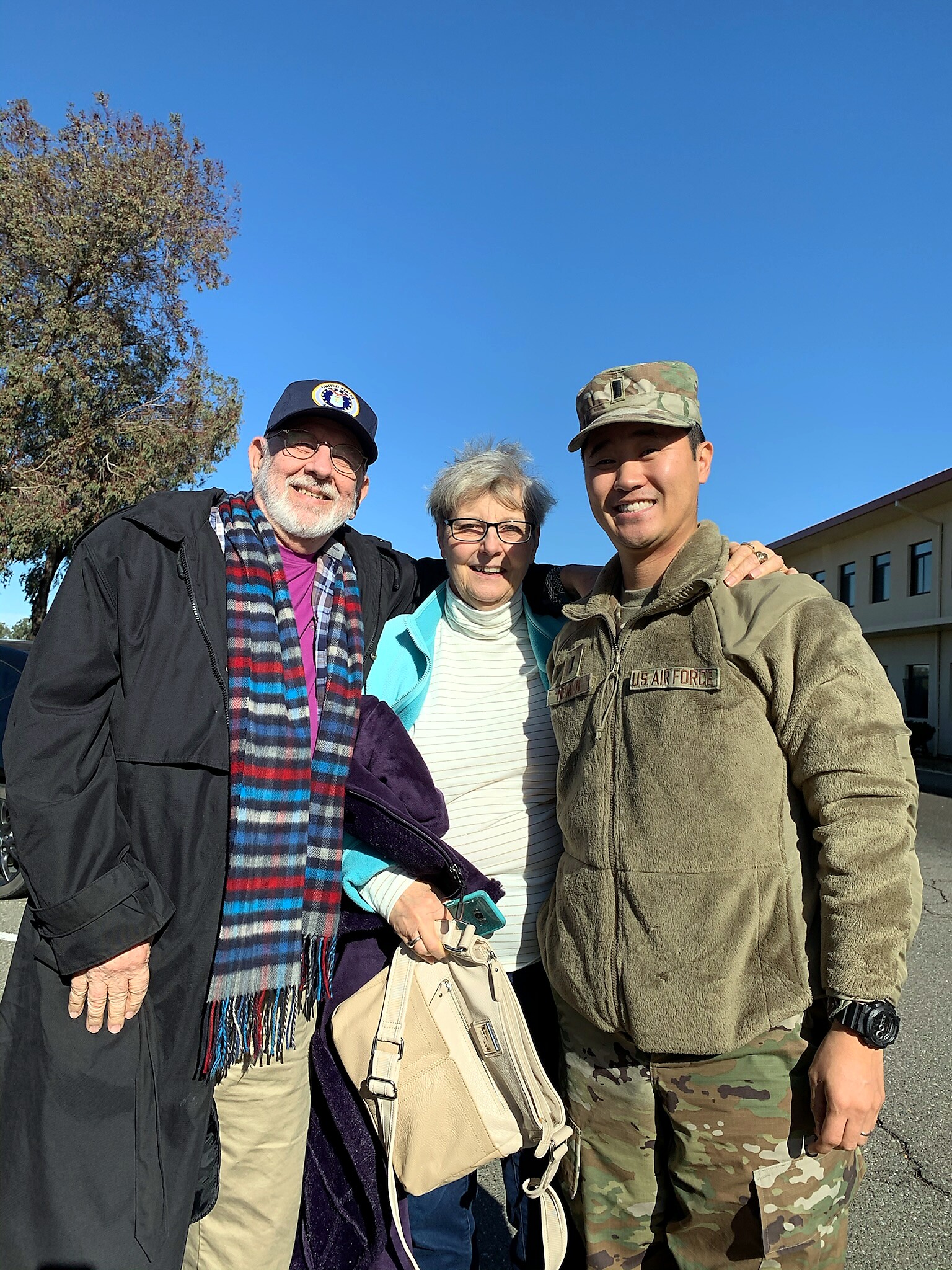 Three smiling adults posing together outside on a sunny day: a man in a black jacket left, a woman in a turquoise jacket center, and a man in a U.S. Air Force camouflage uniform right.