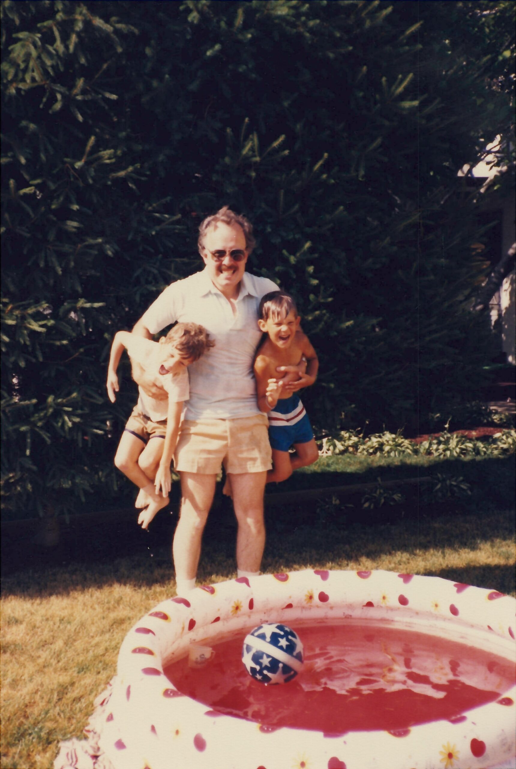 Man wearing sunglasses lifts two laughing children outdoors beside a pink patterned inflatable pool in a backyard.