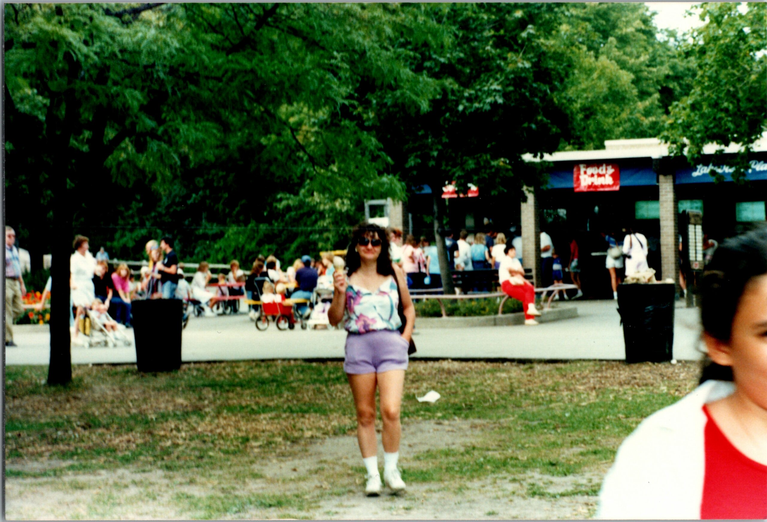 Woman in pastel shorts and sunglasses walks toward camera in a busy park festival with people sitting at tables and food stalls in the background.