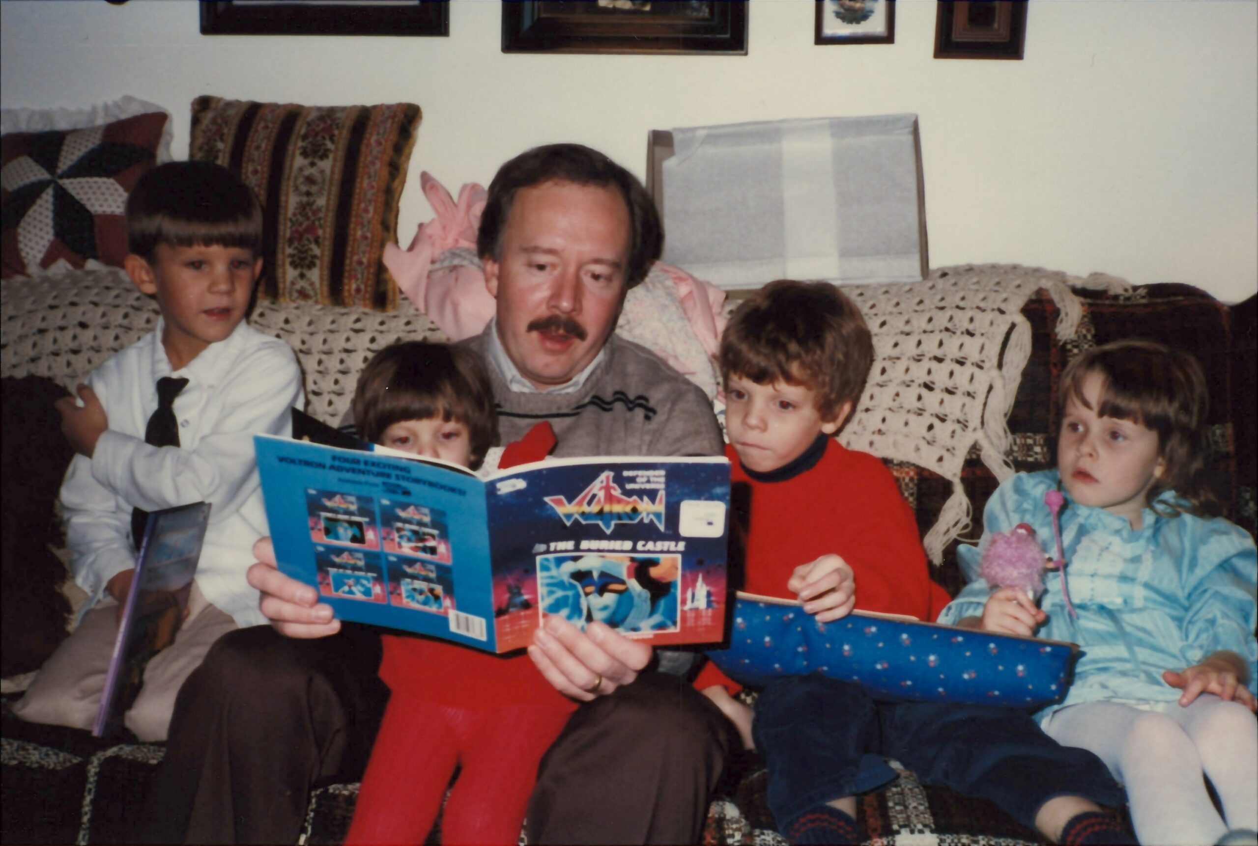 Man with a mustache reading a colorful book to four children on a patterned couch in a living room.