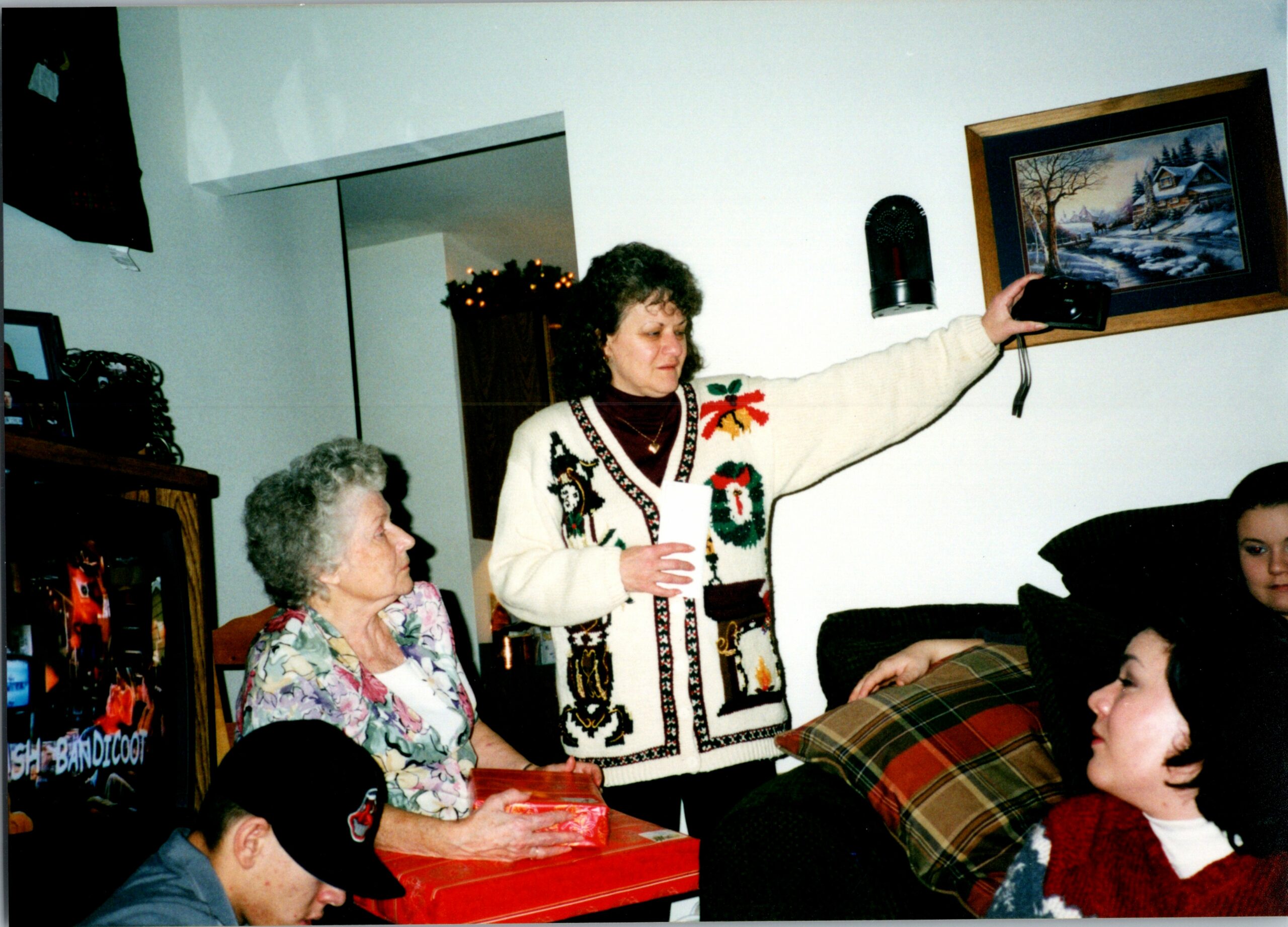 Woman in a festive Christmas sweater extends an arm and hands a wrapped gift to a child during a holiday gathering in a living room.