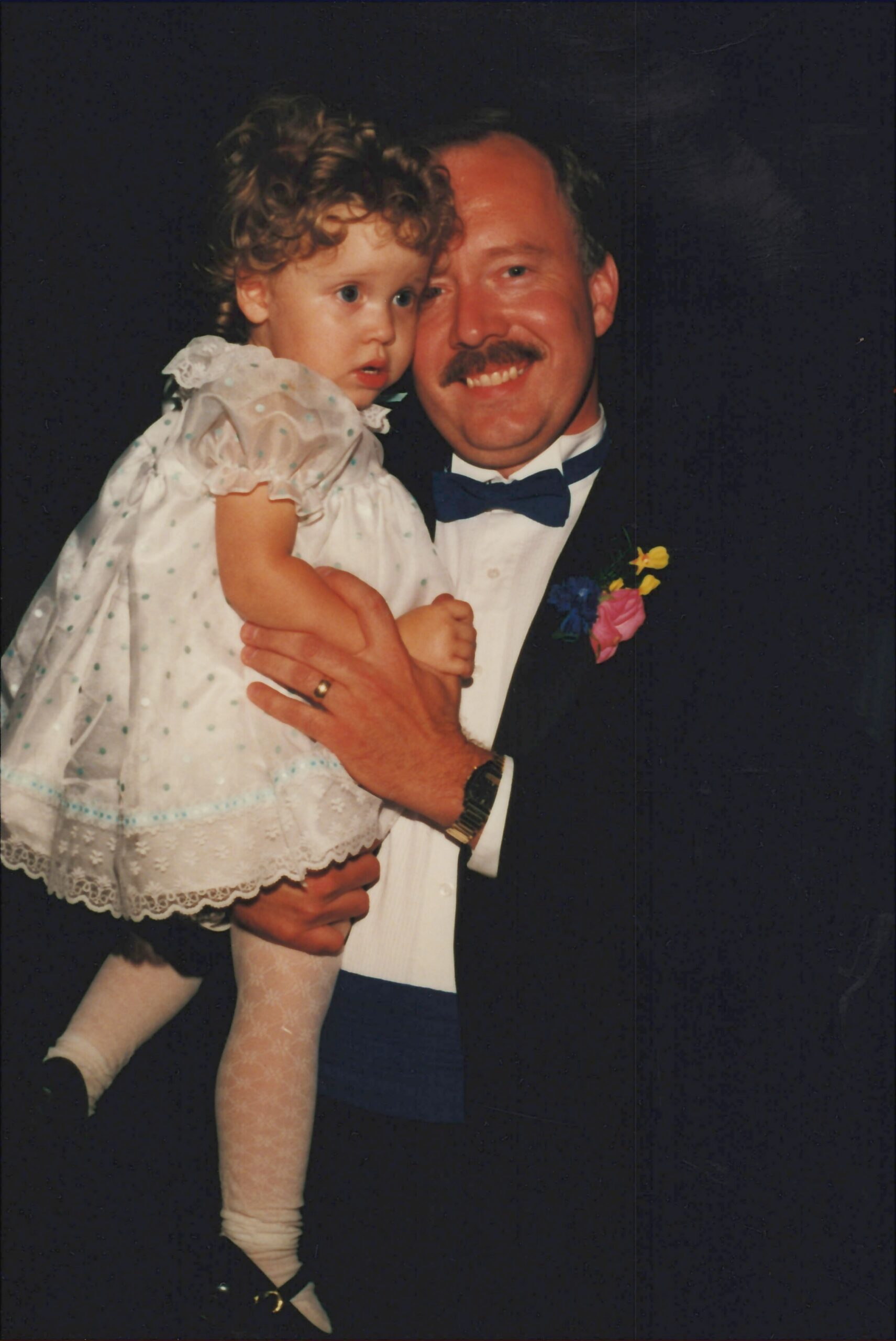 Man in a tuxedo holding a young girl in a white lace dress at a formal event, both looking toward the camera with smiles.