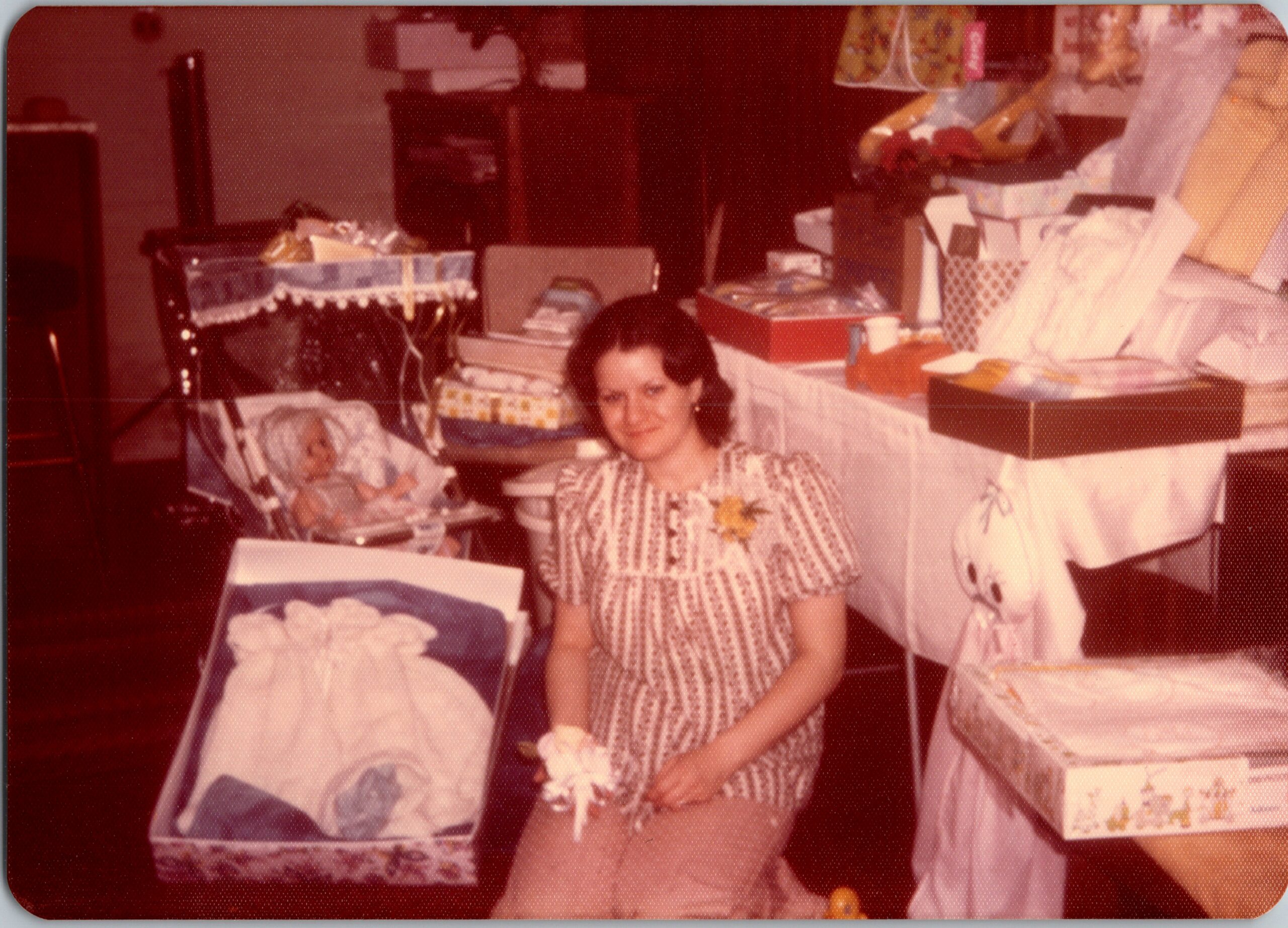 Pregnant woman in a patterned dress stands beside a bassinet filled with blankets, surrounded by gifts at a baby shower setting.
