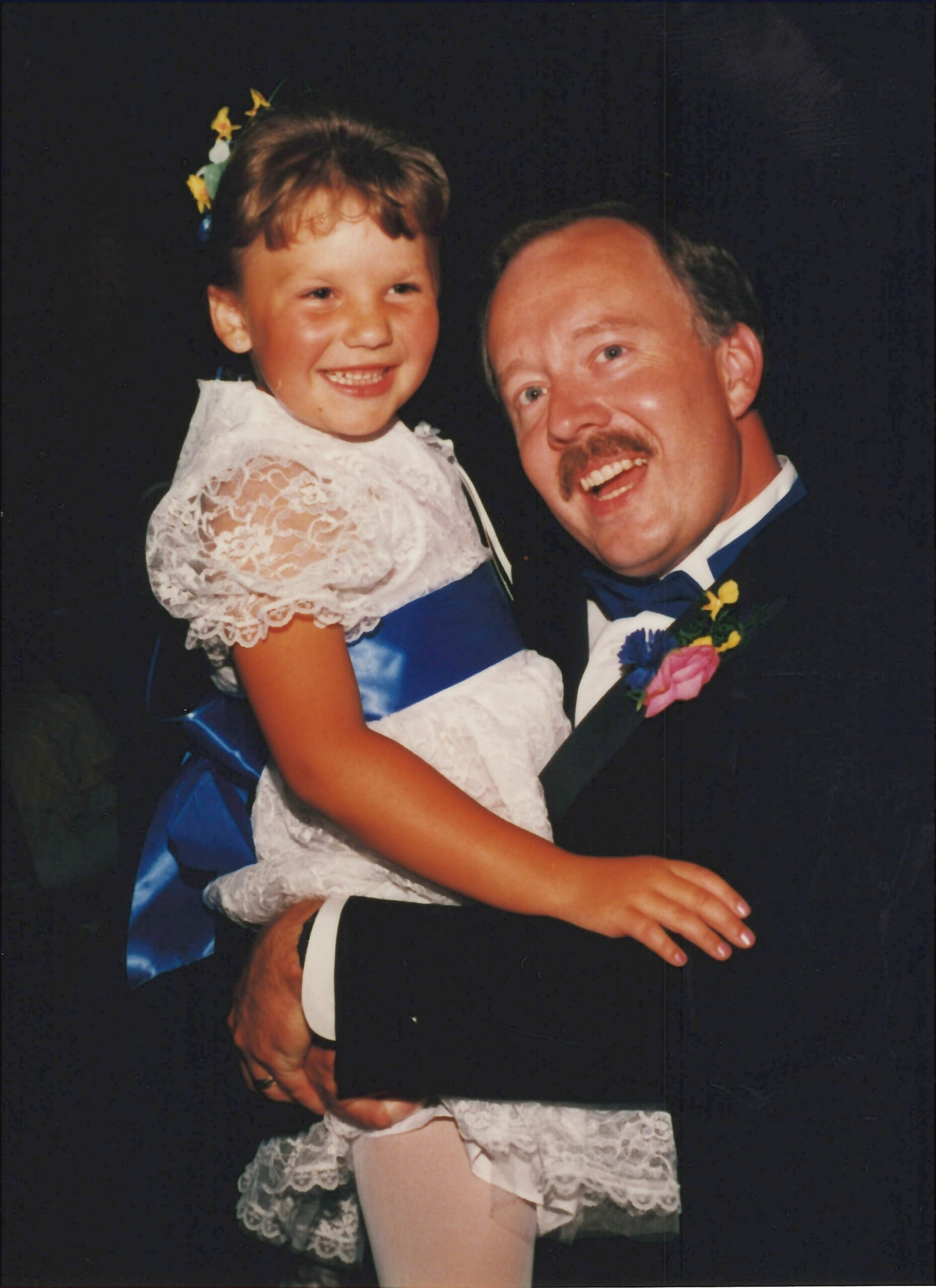 A smiling man in a tuxedo holds a young girl in a white lace dress with a blue sash at a formal event.